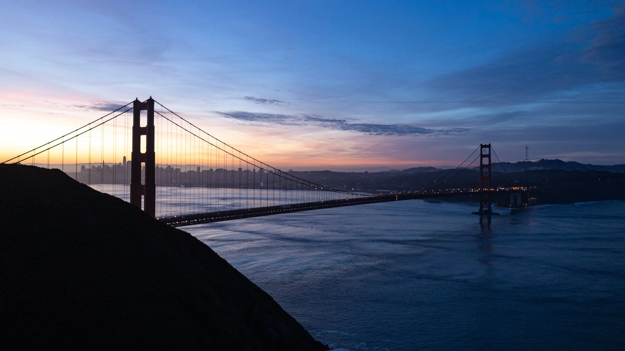The Golden Gate Bridge during sunset with a city skyline in the background, water below, and colorful sky with clouds.