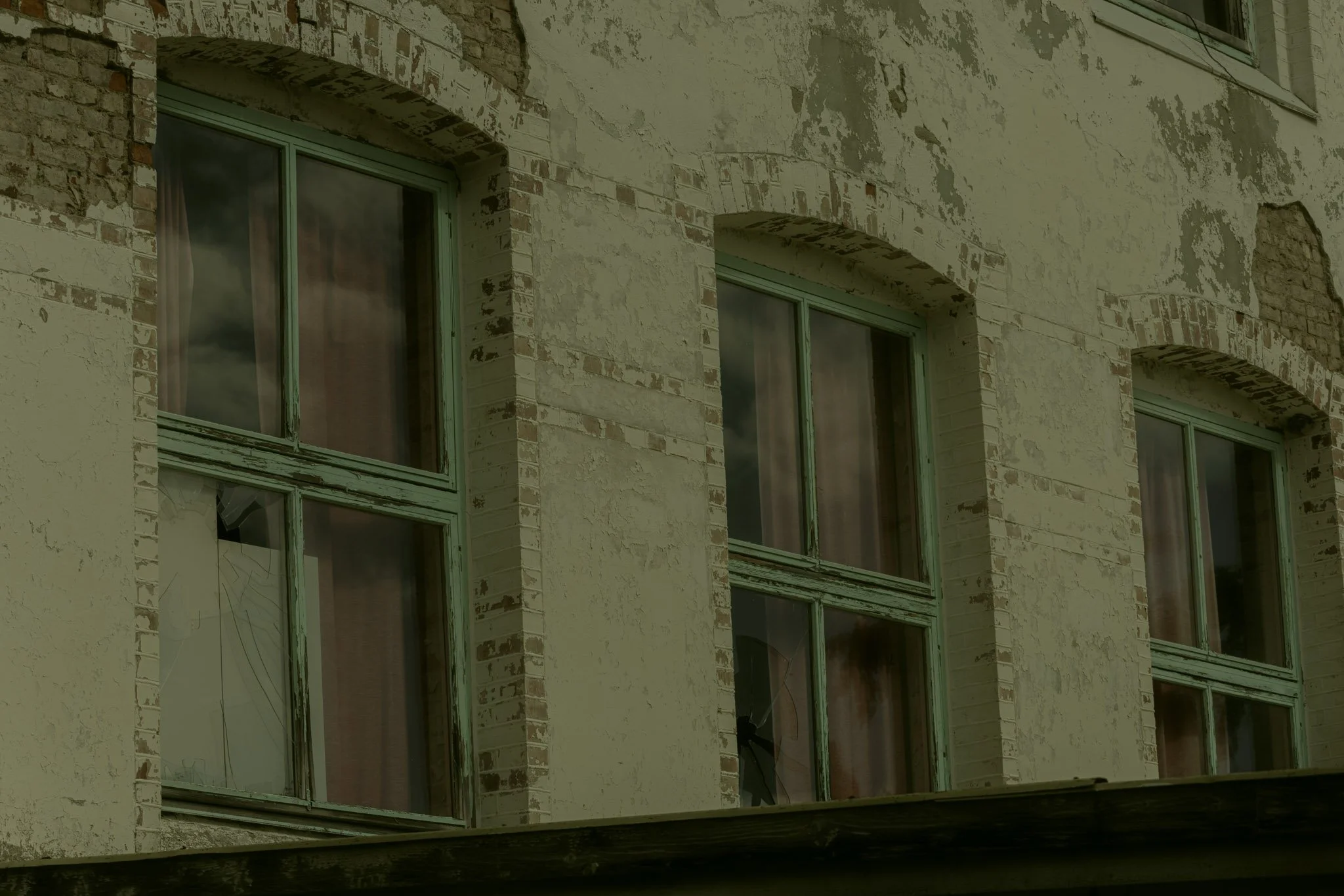 Close-up of an old building's exterior showing broken and damaged windows, peeling paint, and weathered brick and plaster walls.