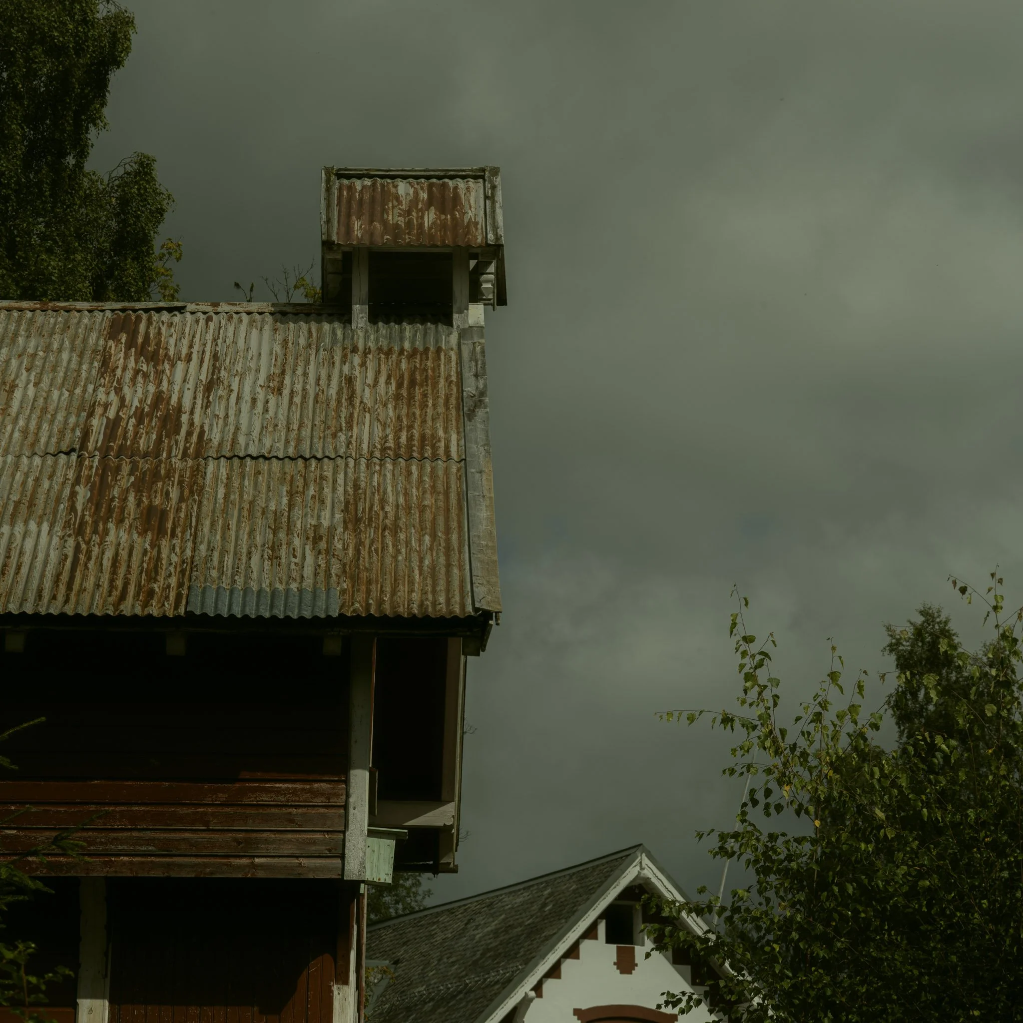 A tall, rusty, corrugated metal structure or shed with a small, elevated section on top, against a cloudy sky, with trees and neighboring houses in the background.