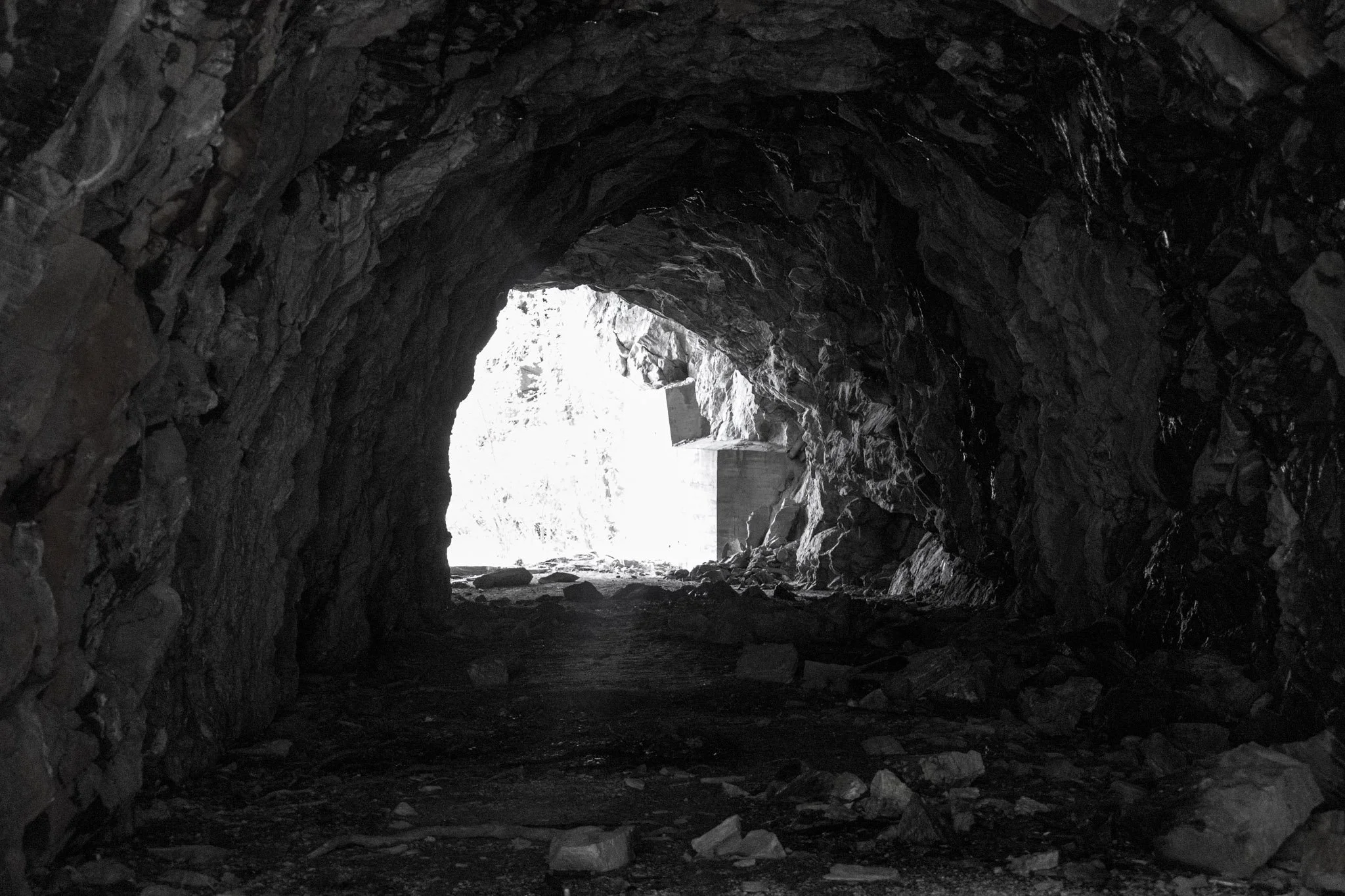 View inside a dark, rocky tunnel with light visible at the far end, revealing rocks and debris along the tunnel floor.
