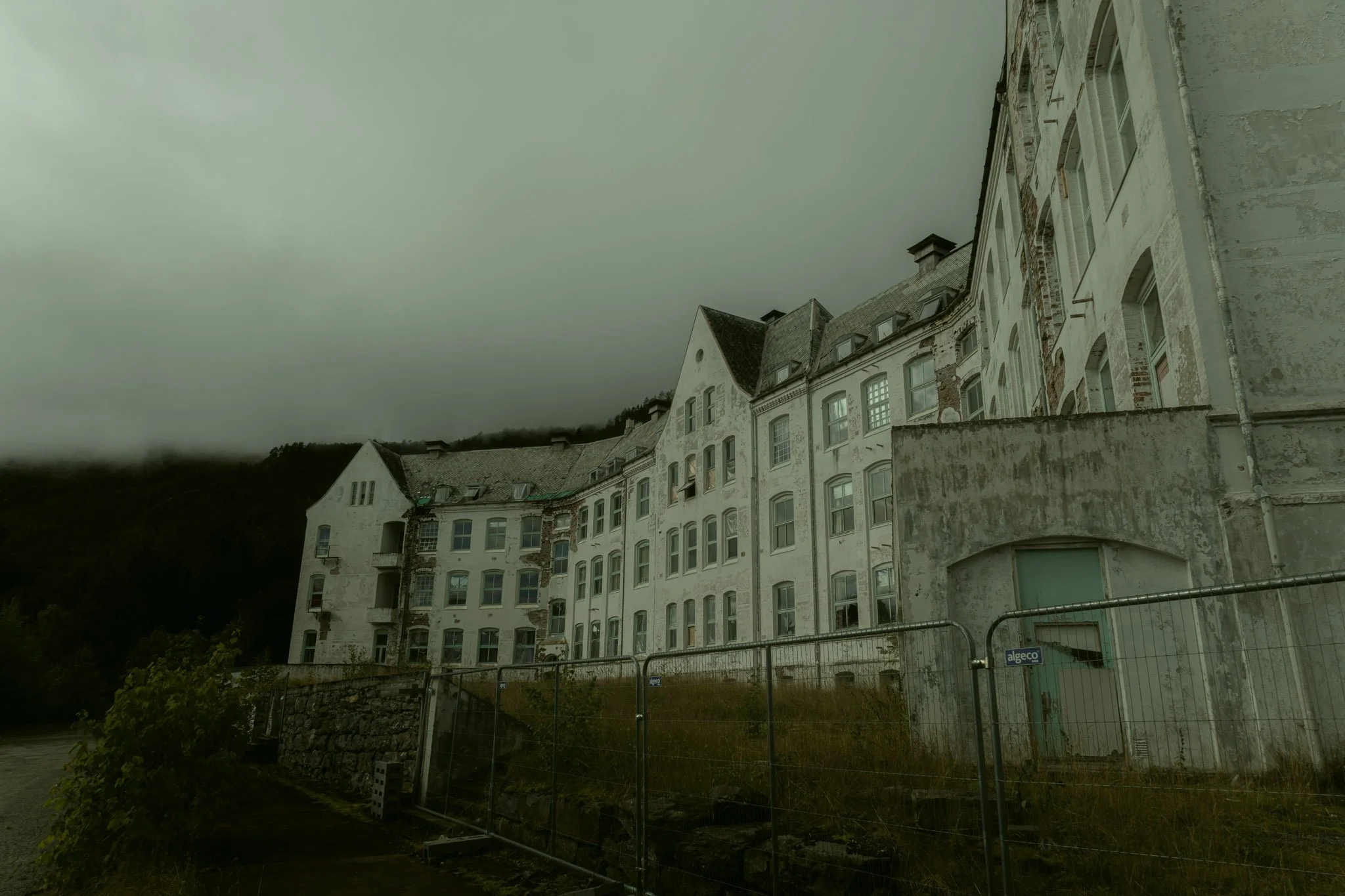 A row of old, white multi-story buildings with arched windows and brick accents, standing on a hillside under a cloudy sky, with a construction fence and greenery in the foreground.