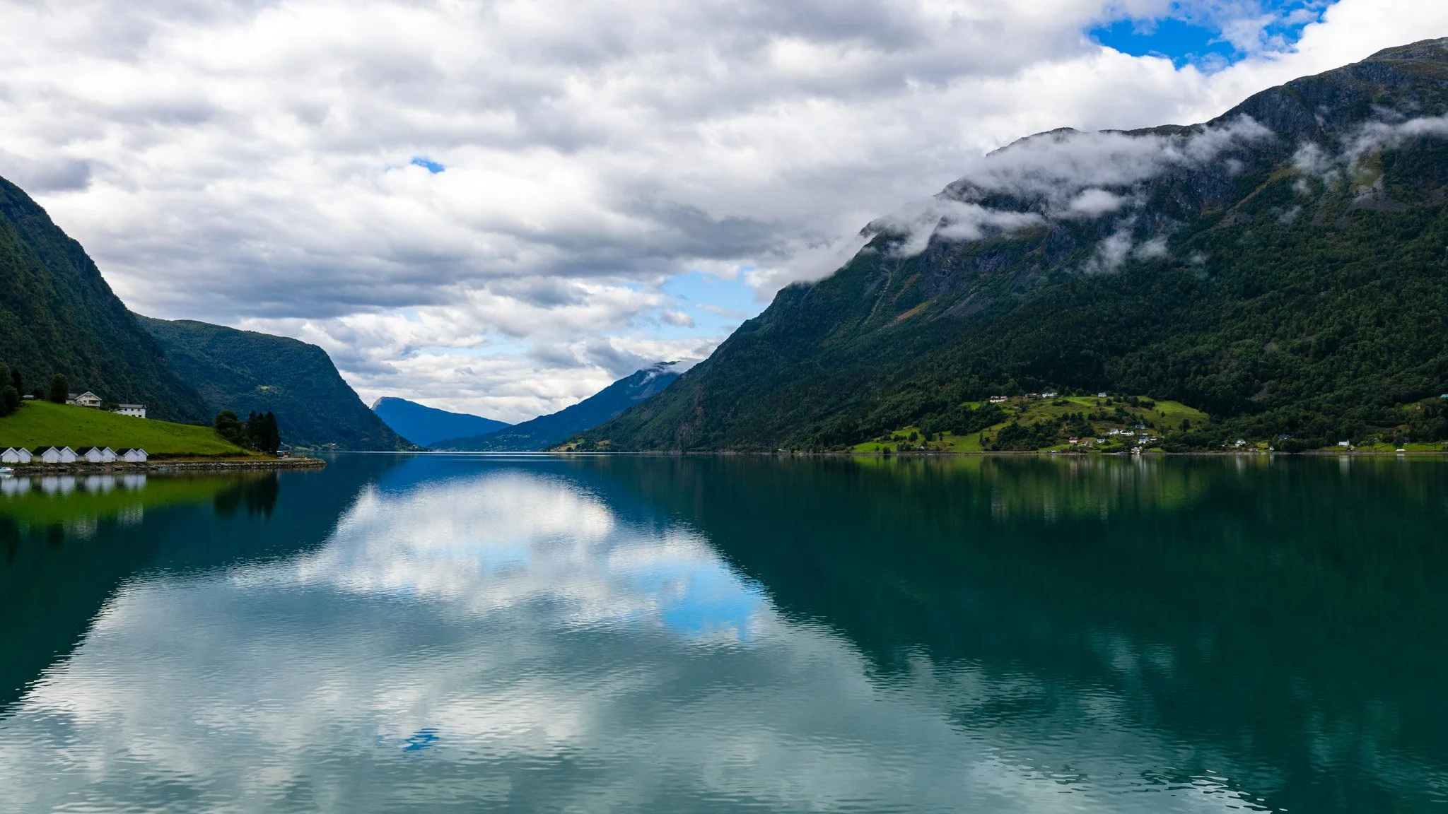 A scenic view of a calm body of water surrounded by green mountains with some clouds and patches of blue sky above.