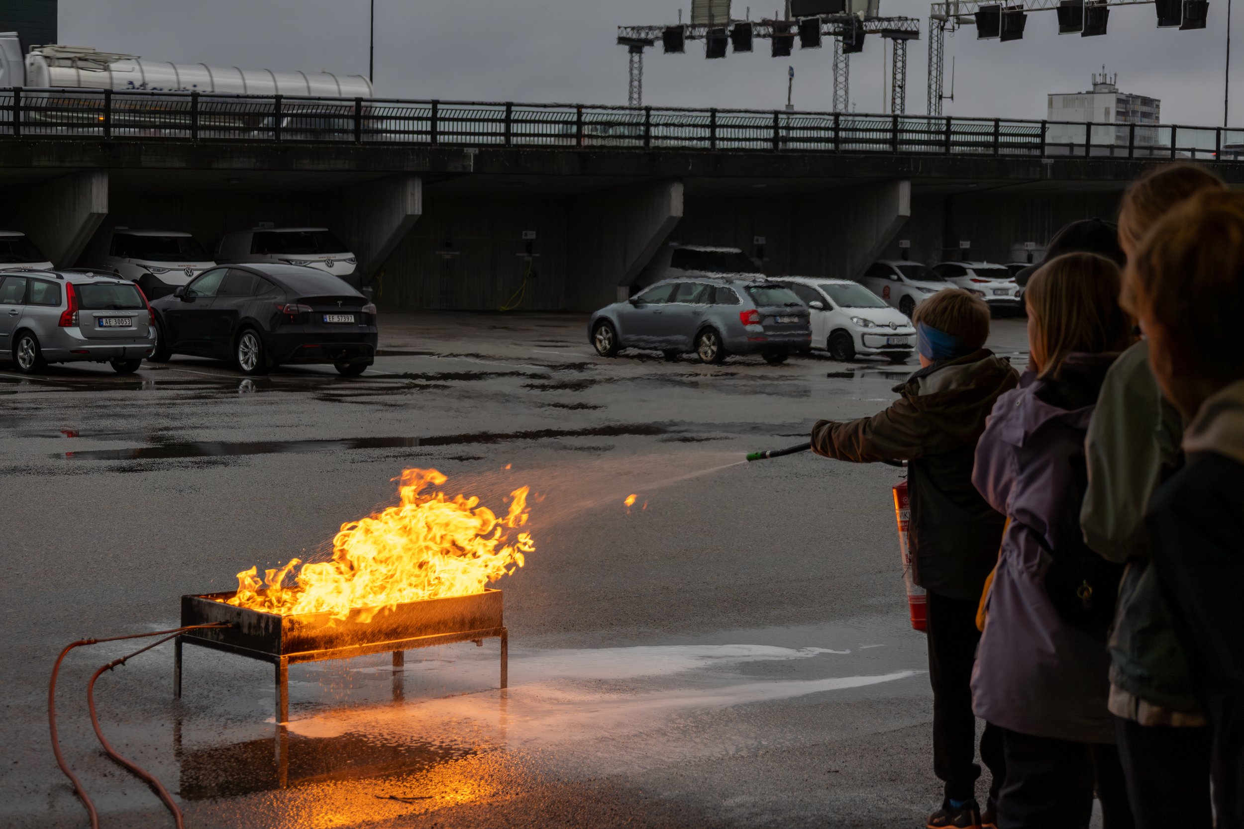 Children standing near a fire in a parking lot on a rainy day, with parked cars, a bridge, and buildings in the background.
