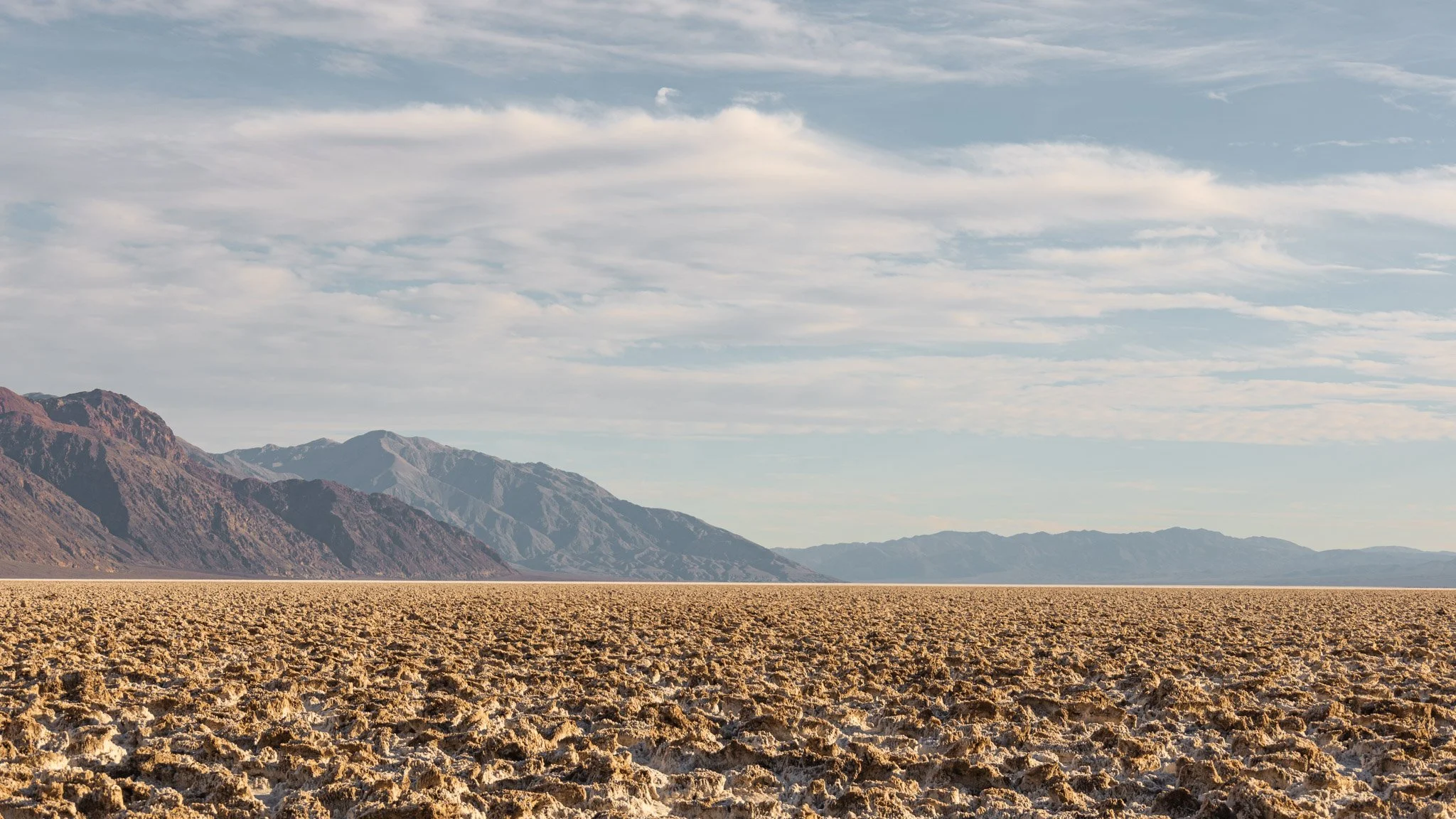 A vast dry desert terrain with cracked, baked earth, mountains in the distance, and a partly cloudy sky above.