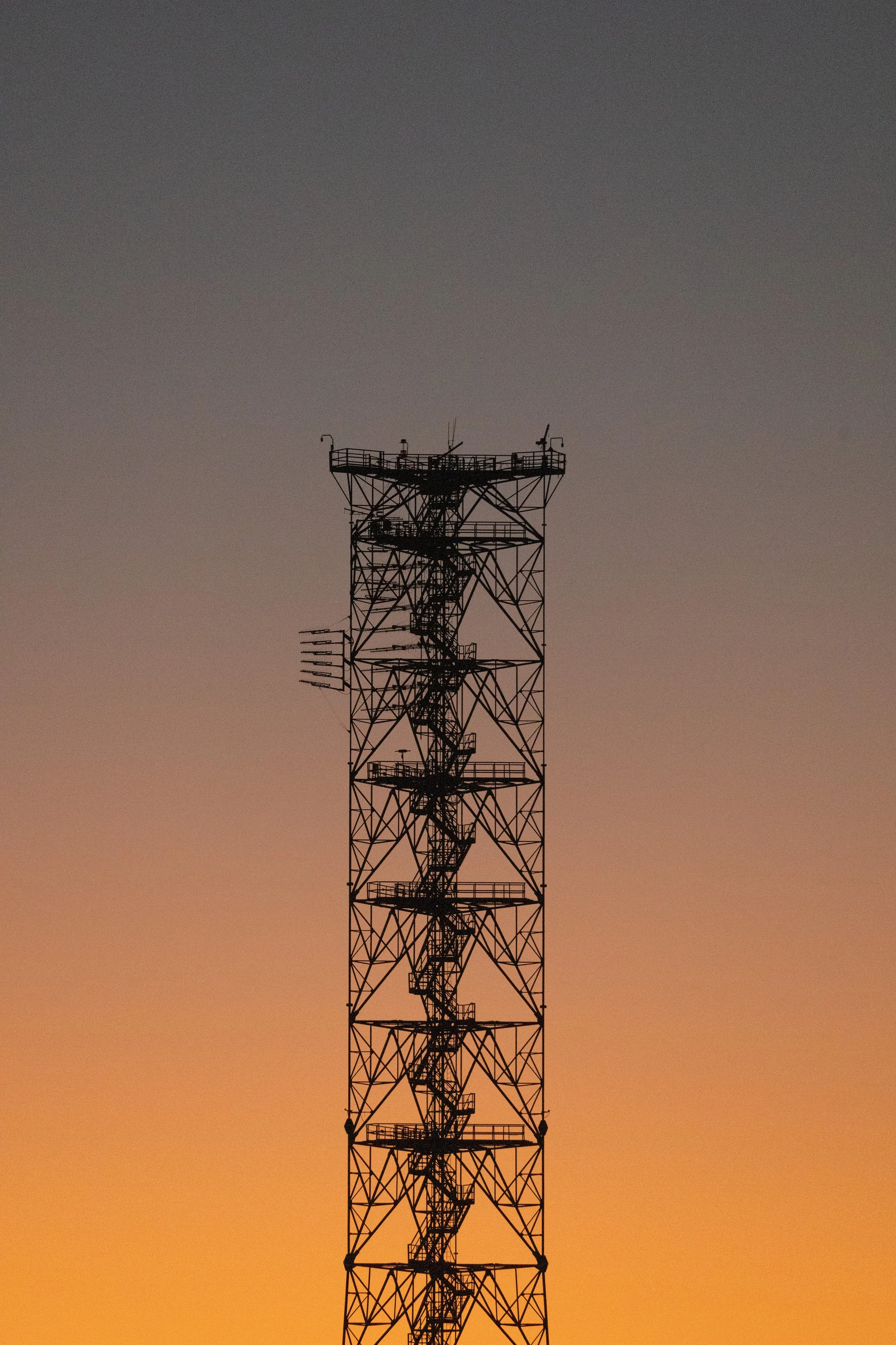 A tall, black metal radio or communication tower with antennas and platforms at different levels, set against a gradient sky at dusk with orange and purple hues.