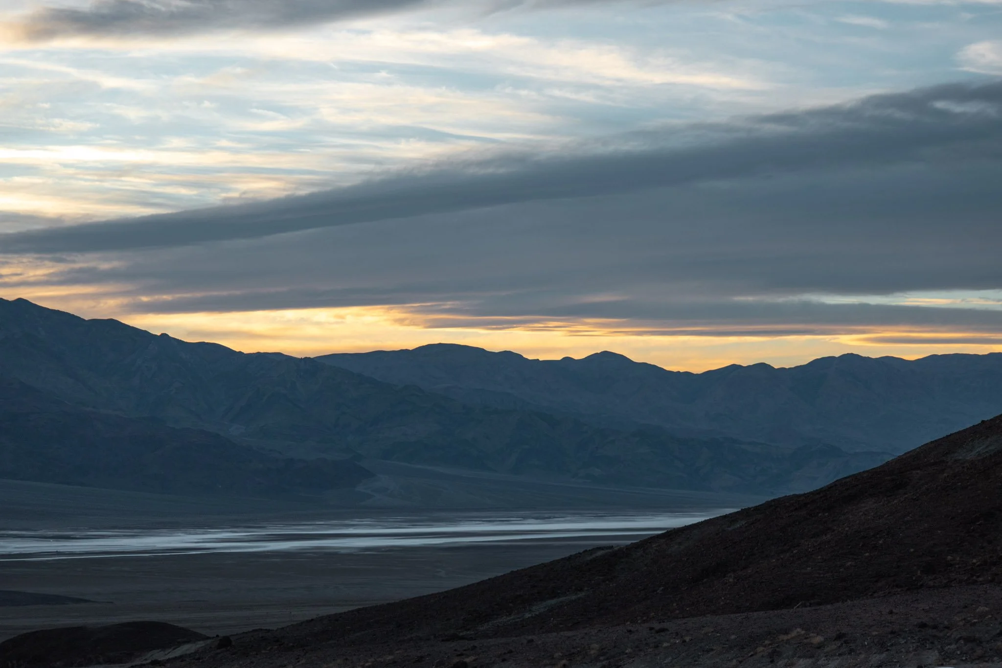 Scenic view of a lake surrounded by mountains at sunset in Death Valley, with a cloudy sky and shadows cast over the landscape.
