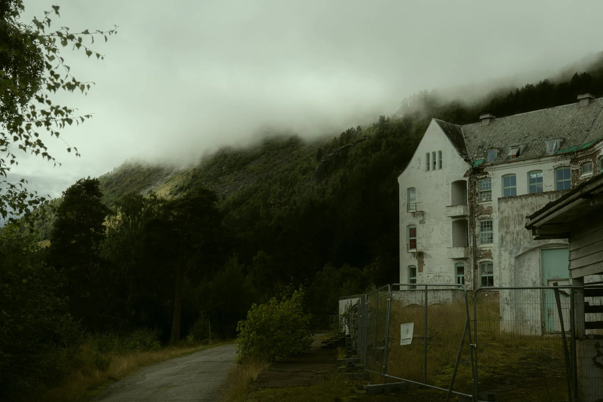 A weathered multi-story white house with a steep roof, located near a forested hillside with low hanging fog, and surrounded by a gravel and dirt pathway with a fenced area.