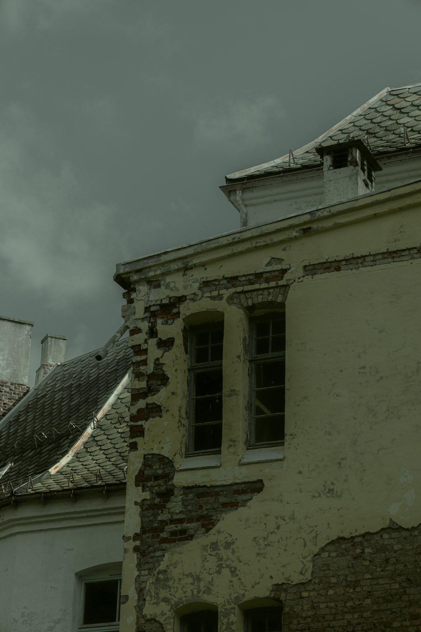 An old, weathered building with peeling yellow paint and exposed brickwork, topped with a shingled roof under a cloudy sky.