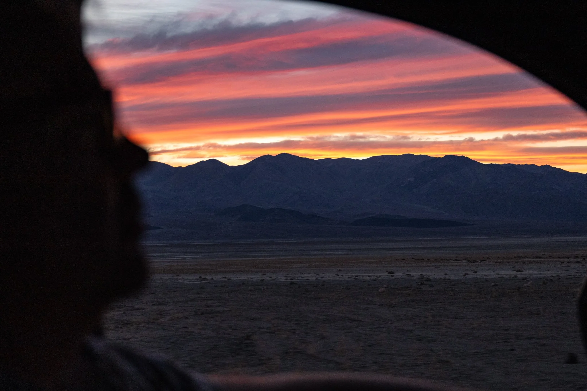 Sunset over mountains in Death Valley seen through a car window with a blurry, dark foreground.