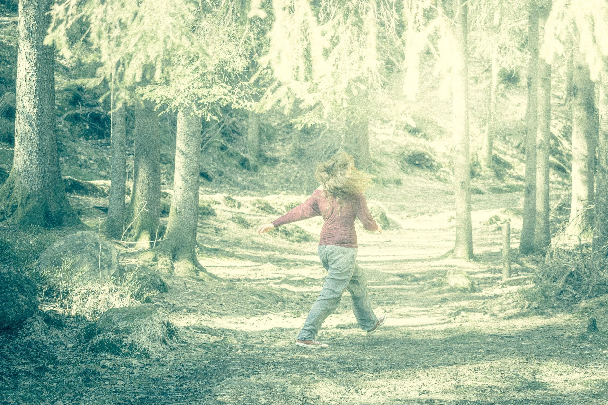 A person with long hair wearing a red jacket and gray pants running through a forest with tall trees and sunlight filtering through the branches.