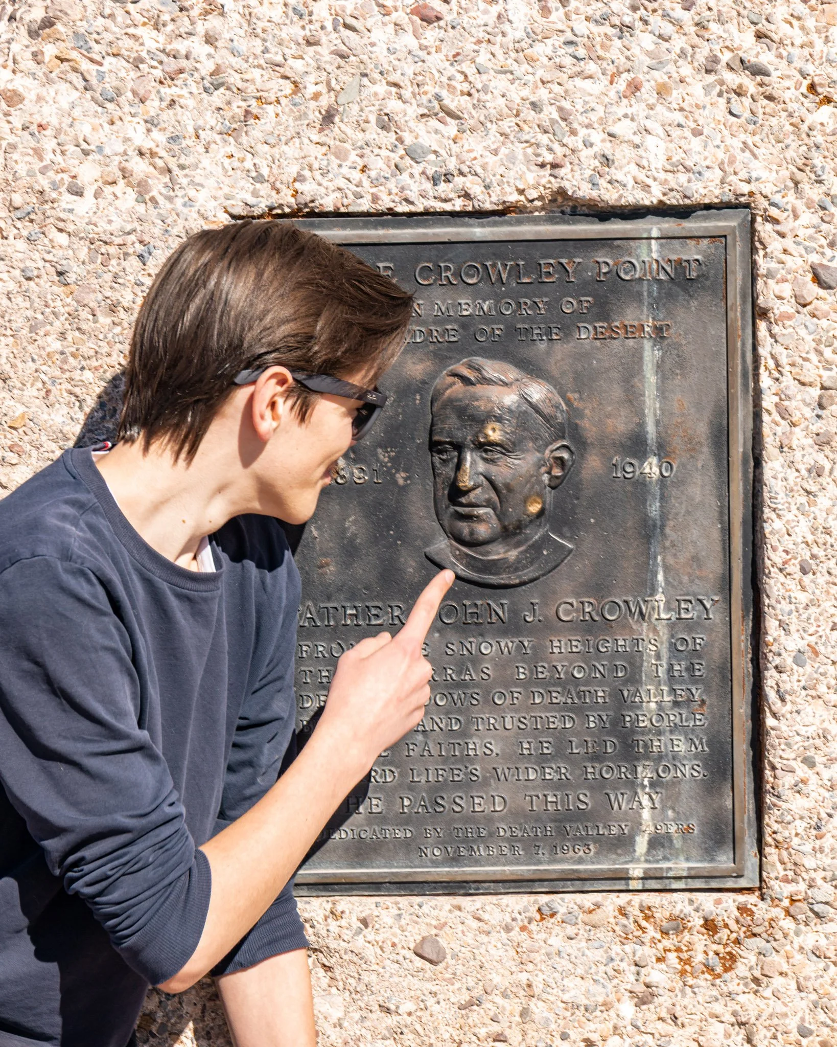 A man with brown hair and sunglasses points at a commemorative bronze plaque mounted on a gravel wall. The plaque honors Father John J. Crowley and commemorates his work starting from 1940.