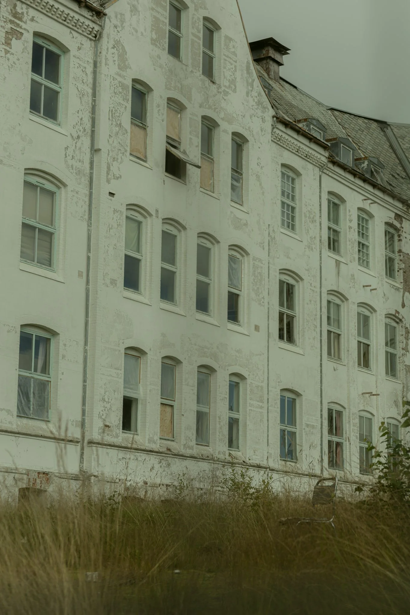 An old, weathered multi-story building with peeling white paint and multiple windows, some boarded up or with broken glass. The building has a steeply sloped roof with dormer windows and appears to be abandoned.