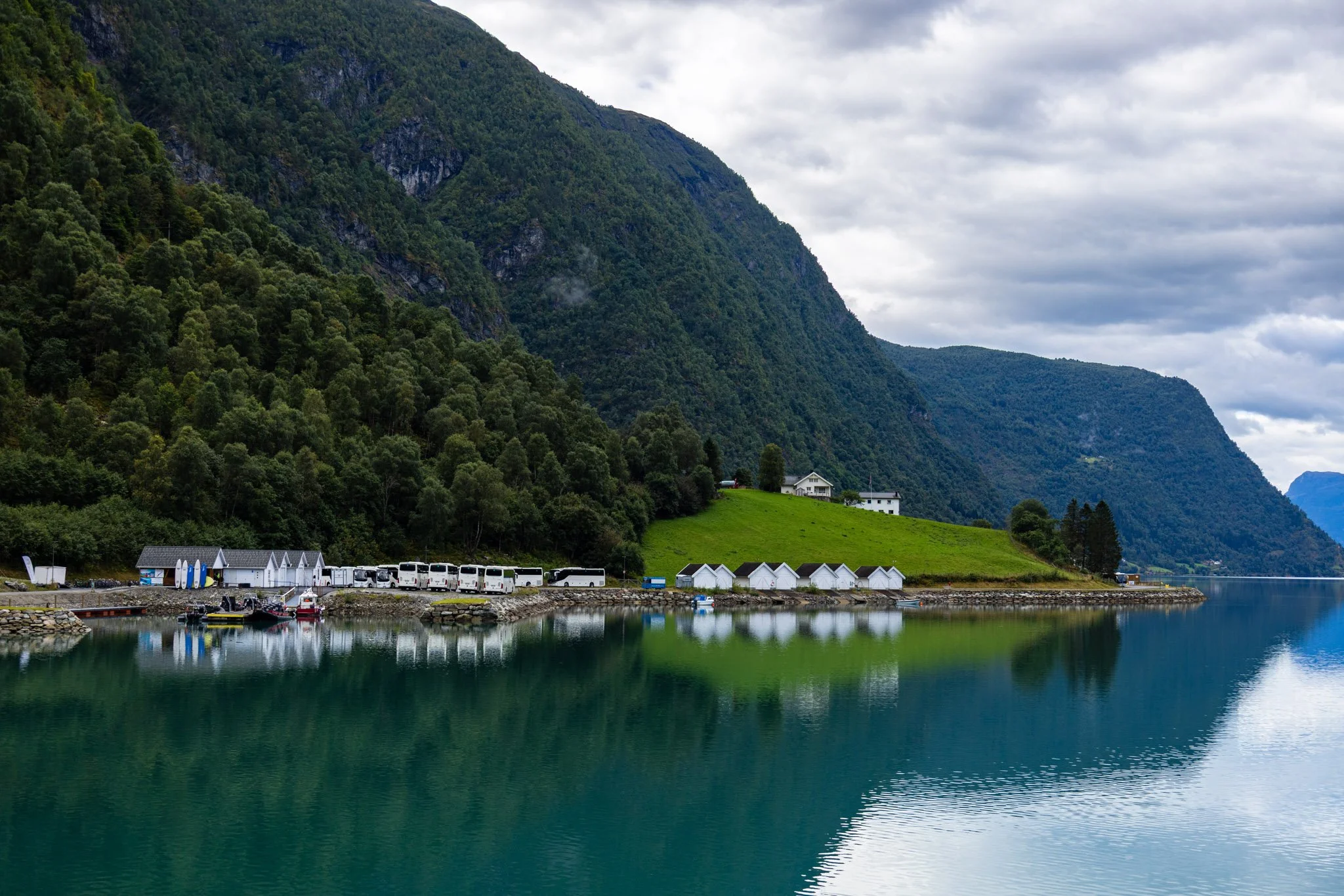 Scenic view of a body of water reflecting a green hillside with small white buildings and a row of parked buses, surrounded by tall green mountains under a cloudy sky.