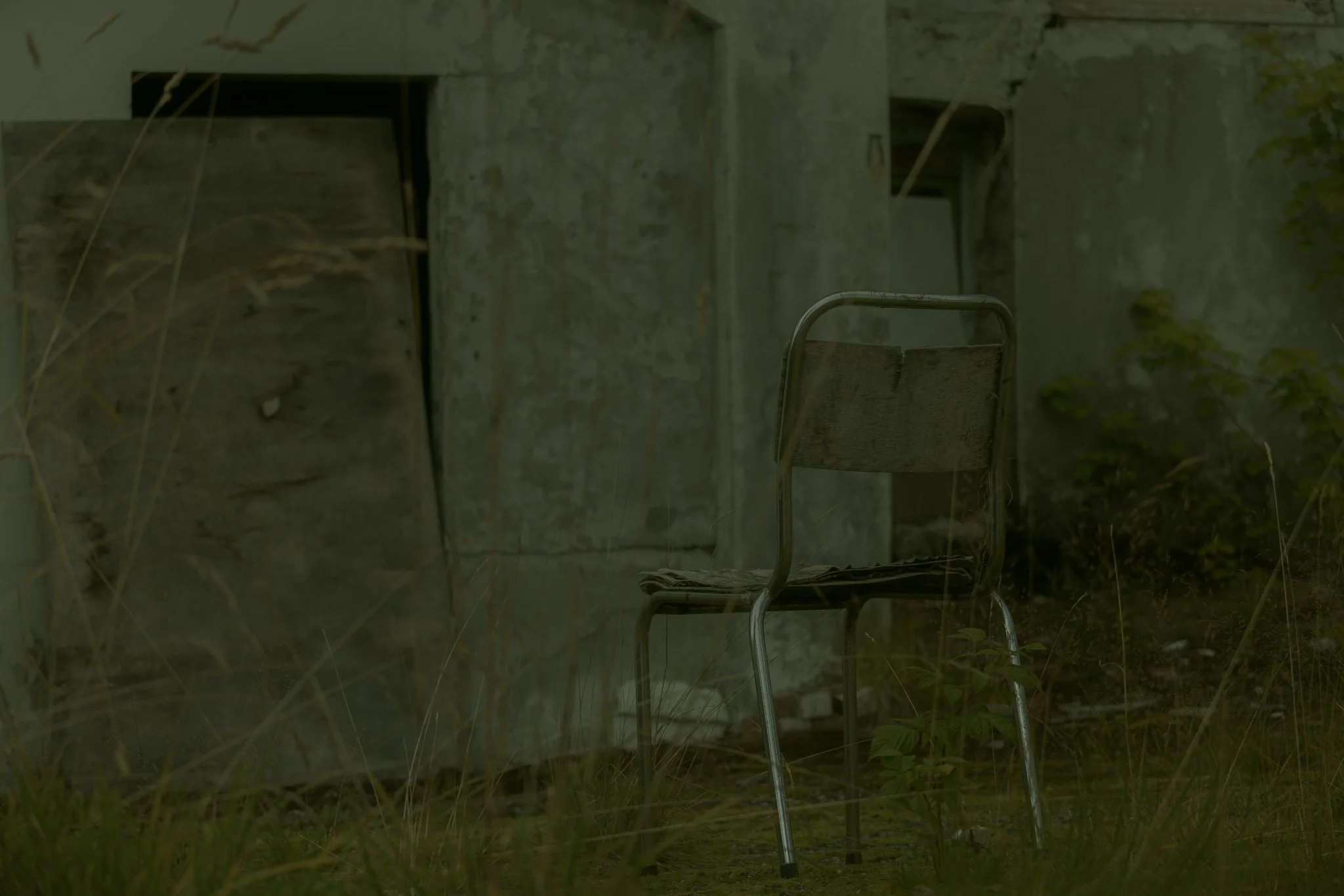 An old, weathered metal chair in front of a concrete or stone wall, with overgrown grass and plants around it.