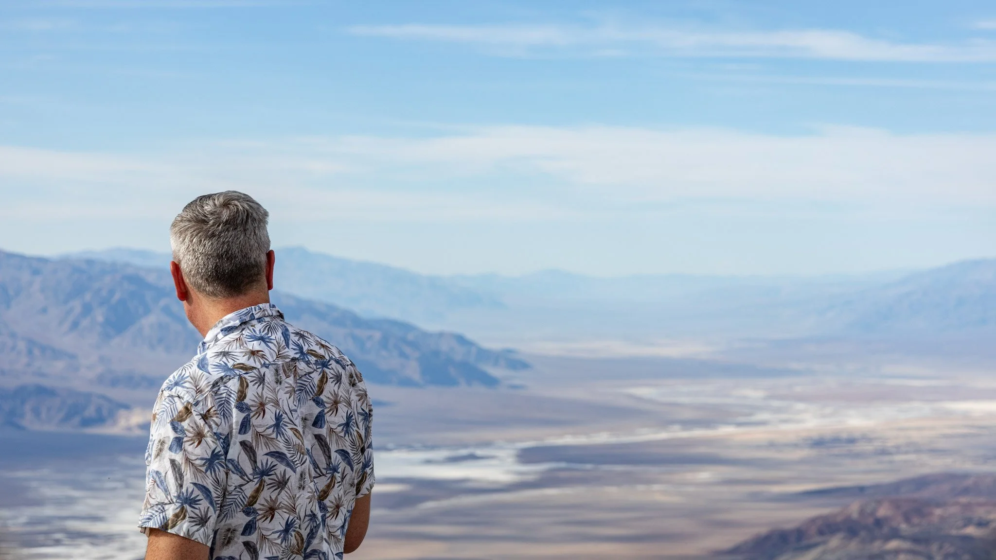 A man with gray hair wearing a short-sleeved shirt with a tropical leaf pattern, standing outdoors and looking towards mountains and a blue sky with wispy clouds.