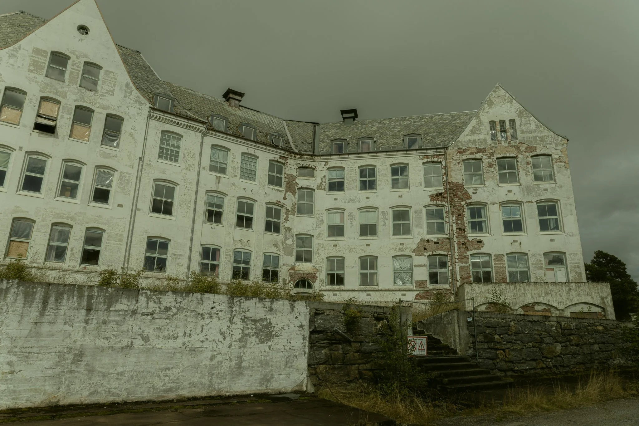 An old, dilapidated white multi-story building with peeling paint and damaged brickwork, featuring multiple windows and a sloped roof under a dark, cloudy sky.
