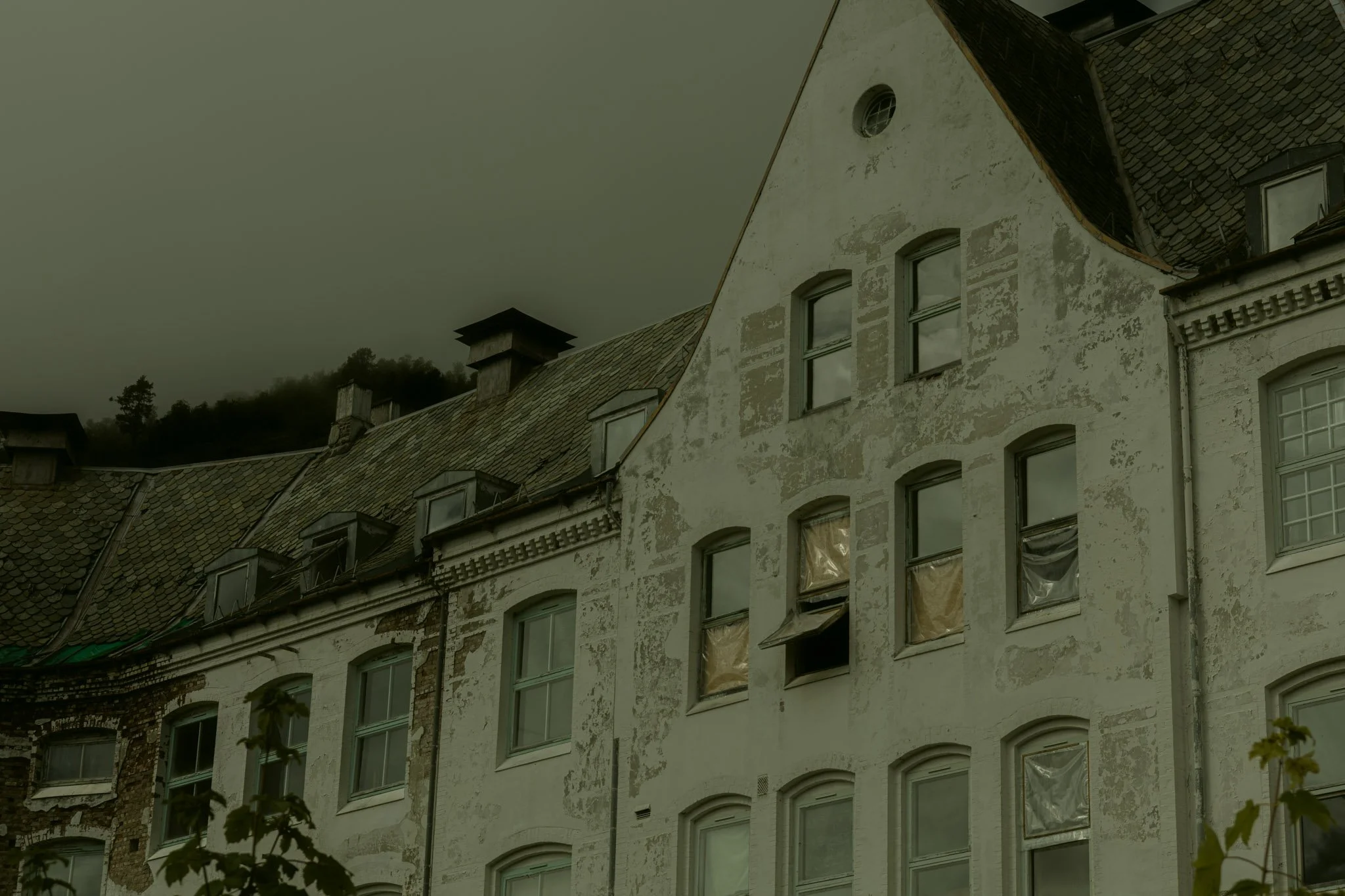 Old multi-story building with peeling white paint, multiple windows, and a tiled roof, under a cloudy sky.