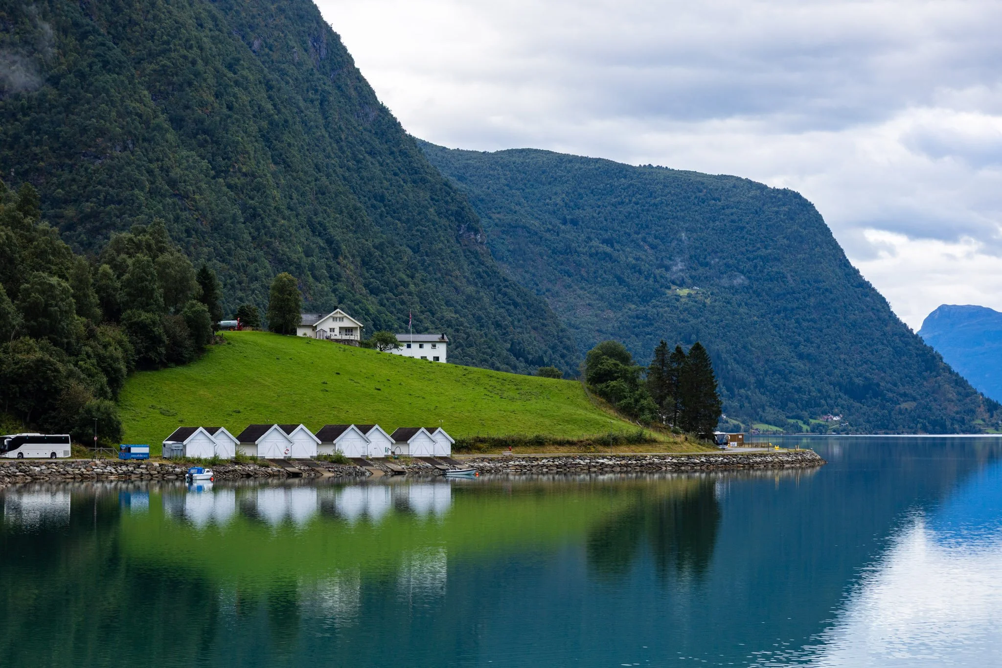Lush green mountains with houses and boats along a calm body of water, under cloudy sky.