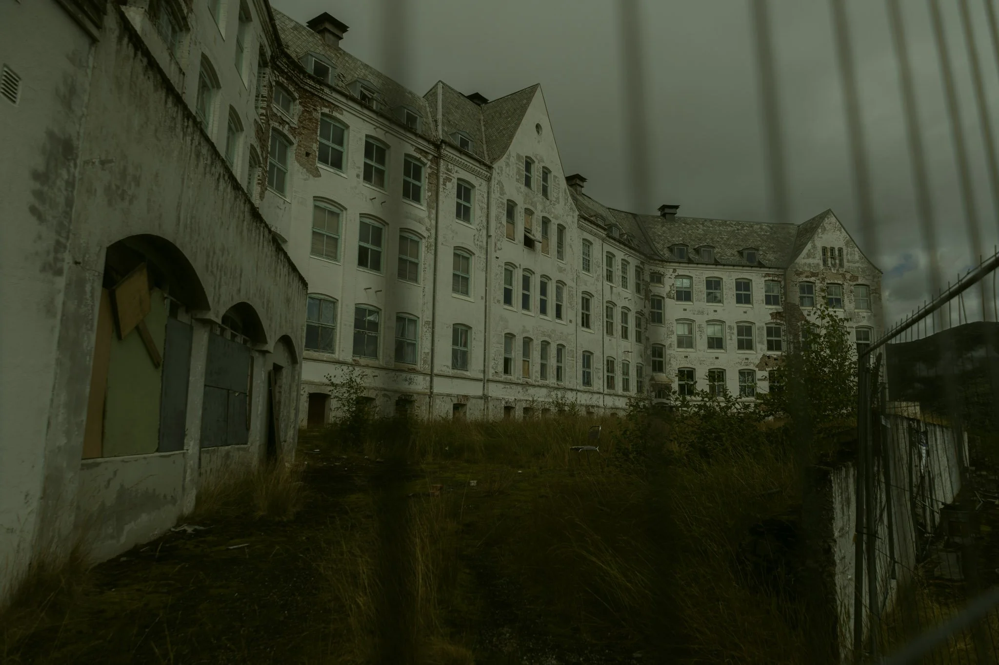 An abandoned, dilapidated building with peeling white paint, broken windows, and overgrown vegetation in the foreground. The building has multiple stories and steep roofs, with a cloudy sky overhead.