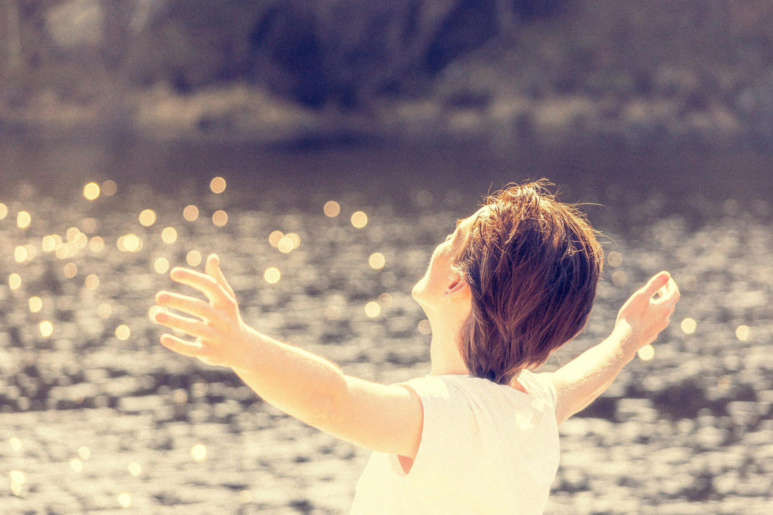 A young woman with reddish-brown hair stands with arms outstretched, facing the water, on a beach during sunset or sunrise, with a bokeh effect of sunlight on the water.