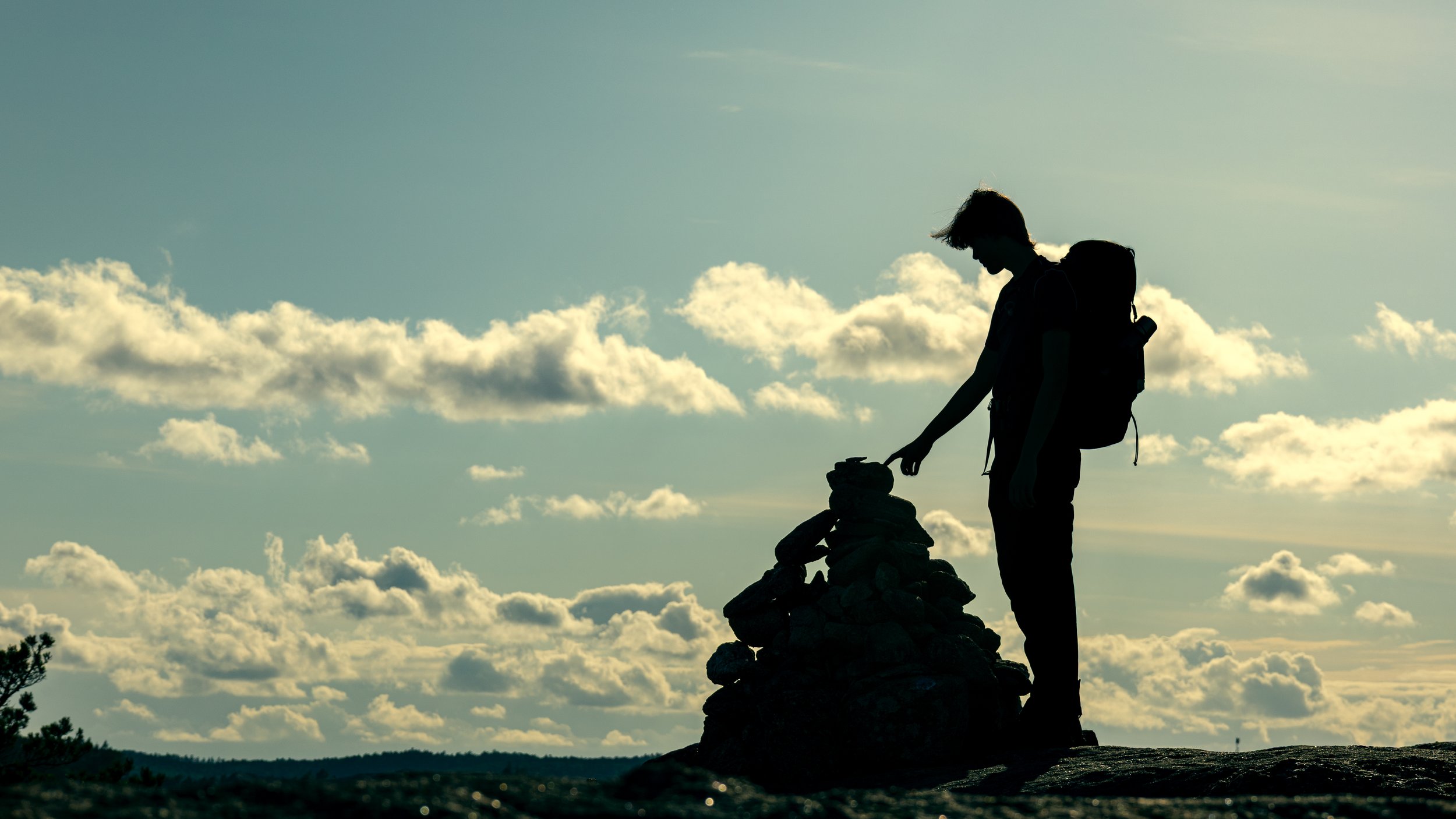 Silhouette of a person with a backpack touching a pile of rocks or cairn on a mountain summit during sunset or sunrise, with a sky filled with scattered clouds.