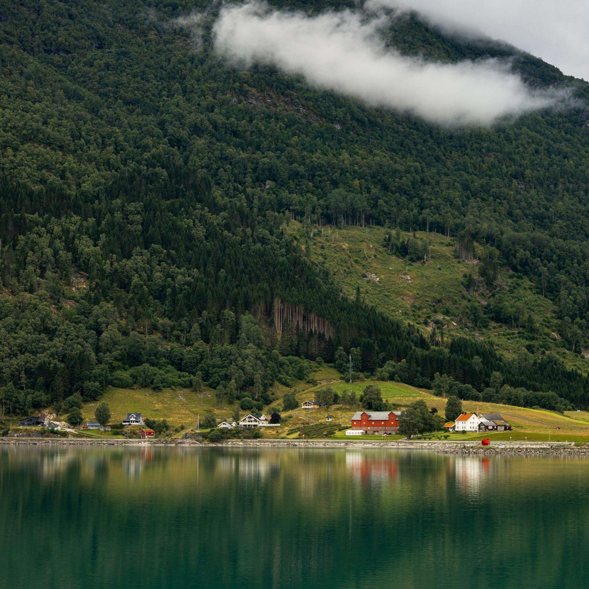 Scenic view of a small coastal town at the base of lush green mountains with low-hanging clouds and a calm body of water reflecting the landscape.