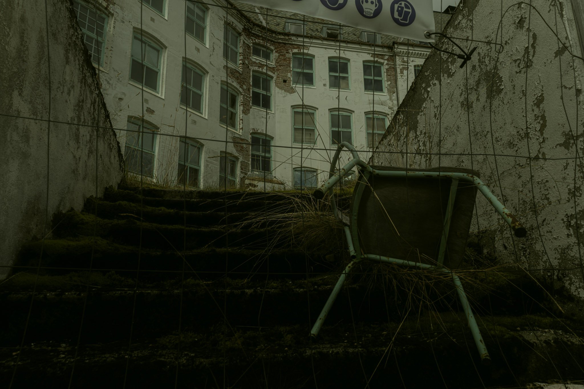View of a dilapidated brick and stucco building through a chain-link fence, with a ground-level trash bin in the foreground, some moss, and dried nettles.