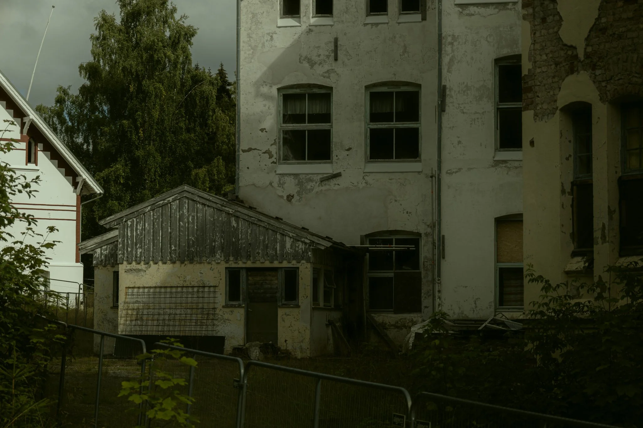 An old, dilapidated building with peeling, dirty white paint and broken or boarded-up windows. There are trees in the background and a small wooden structure in the foreground, surrounded by a fence.