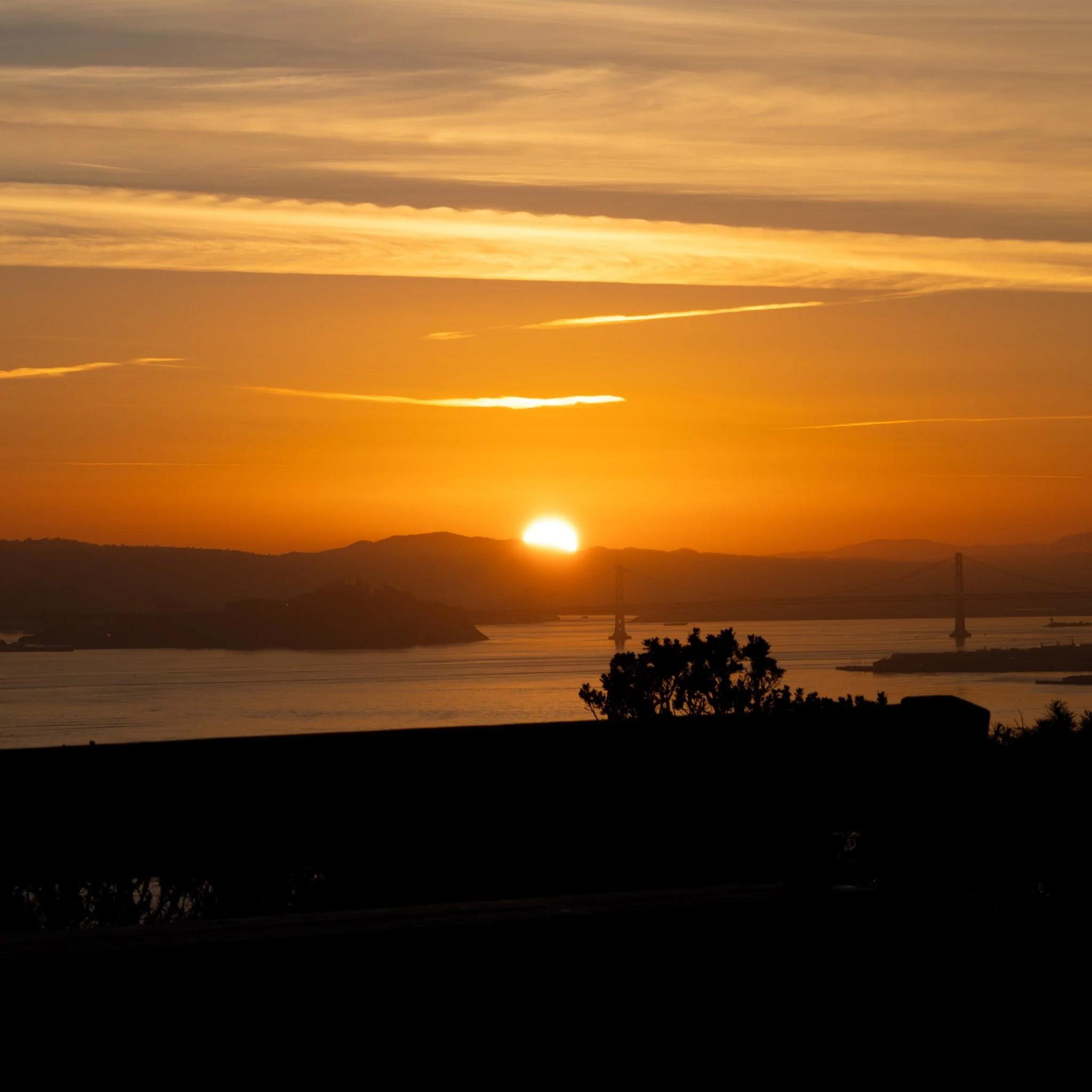 The image shows a scenic view of a sunset over a body of water with distant hills and a bridge. The sky is filled with orange, yellow, and gold hues with scattered clouds.