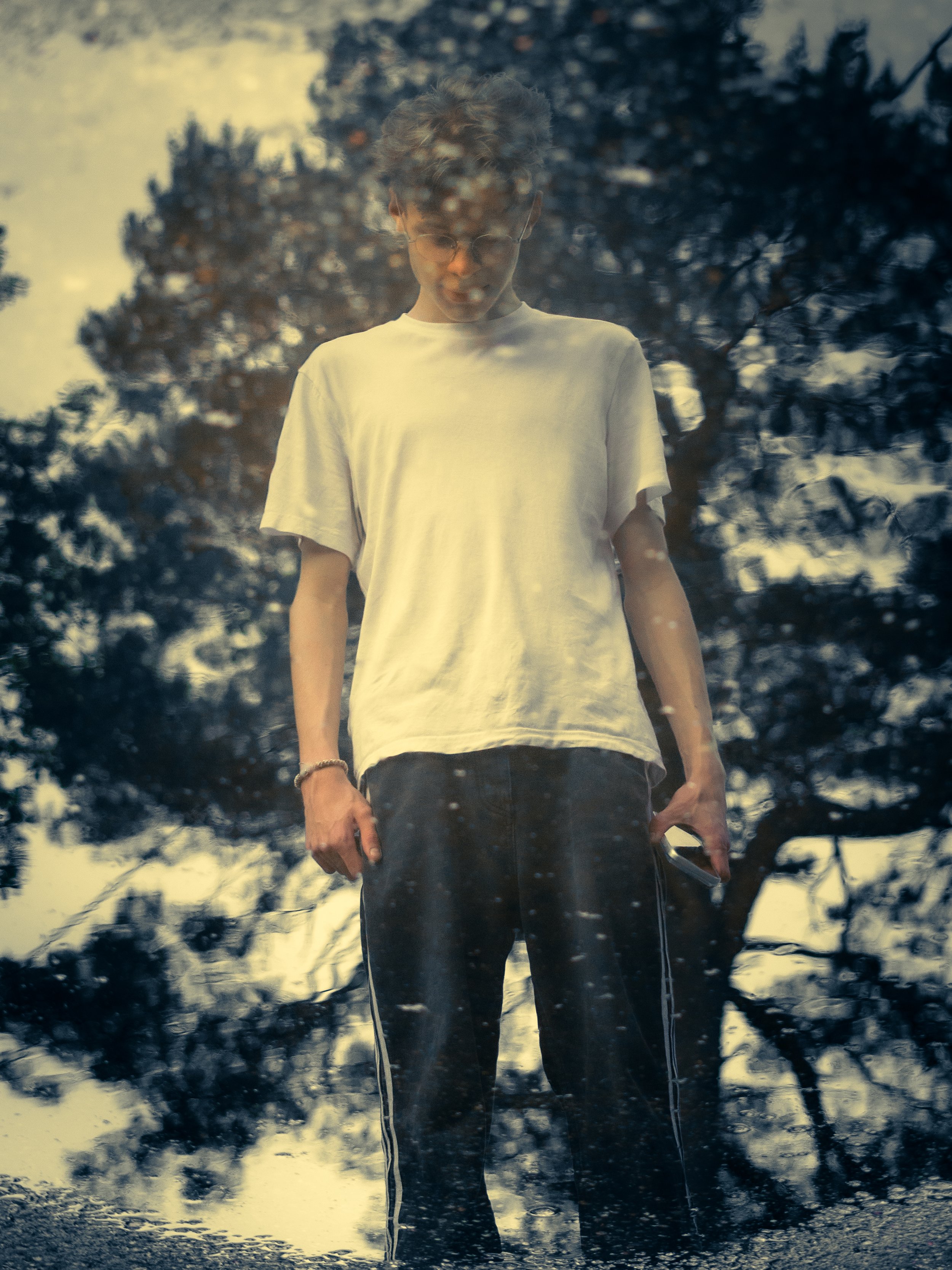 A young man wearing glasses and a white t-shirt looking down, standing outdoors with trees and cloudy sky blurred in the background, viewed through a rain-covered window.