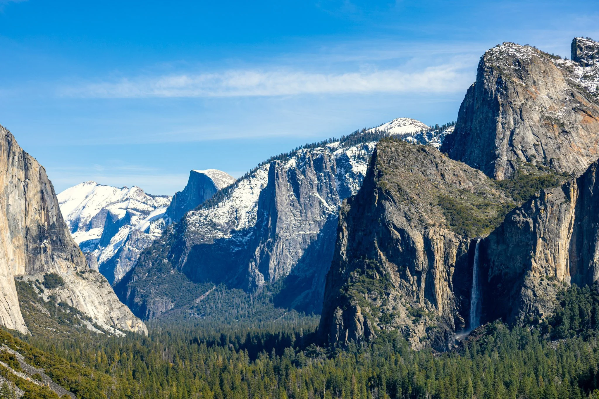 Snow-capped mountains and cliffs under a blue sky with wispy clouds, with a forest at the base.