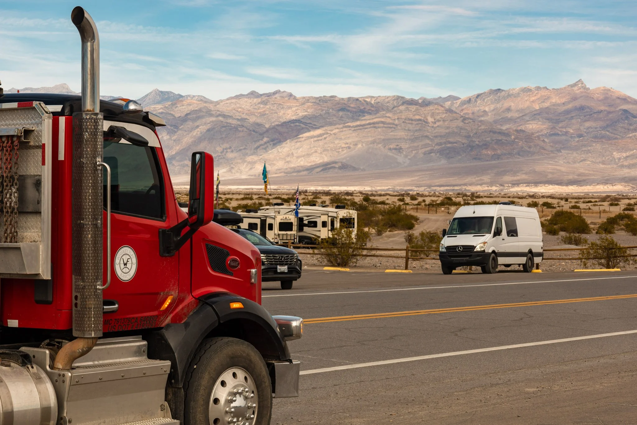 A red fire truck parked beside a white van in a desert parking lot with mountains in the background and blue sky overhead.