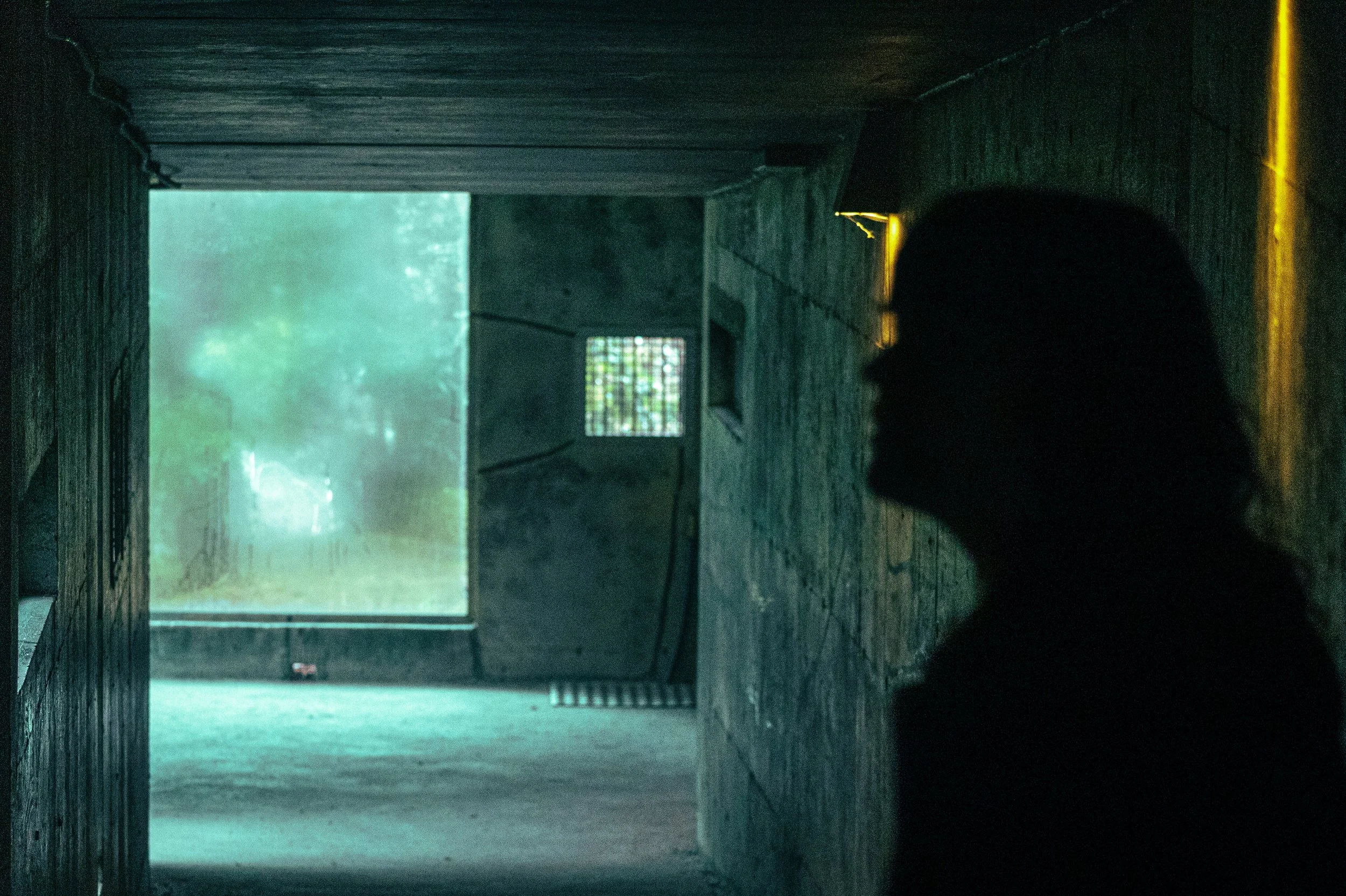 Dark hallway or tunnel leading to an opening with bright light and trees outside. The interior has wooden walls and a small window with bars. The floor appears to be concrete, with some yellow markings near the bottom right corner.