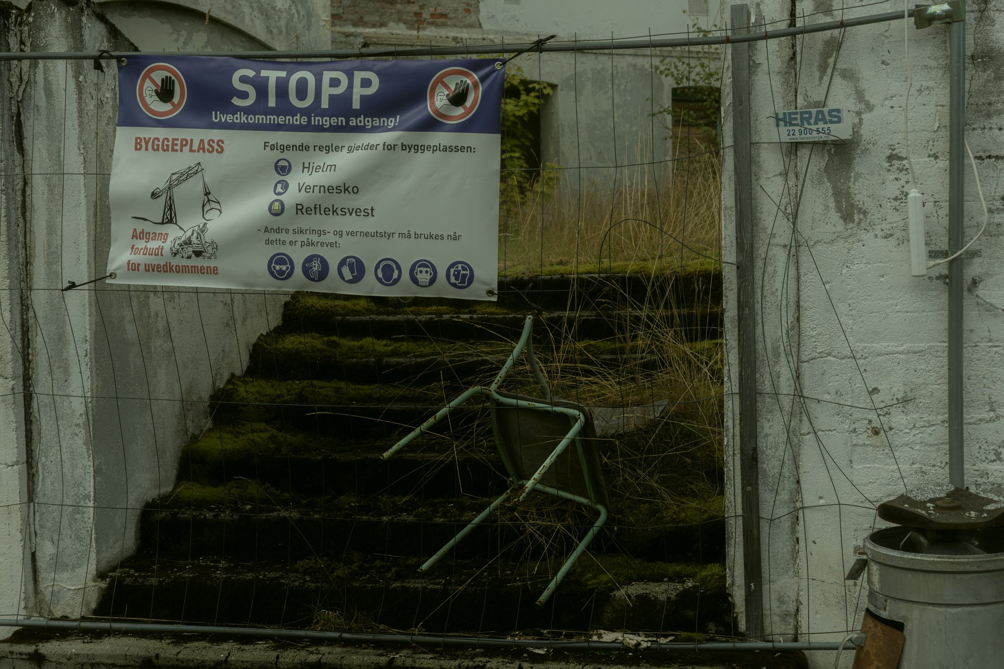 Construction site covered by a wire fence with a sign in Norwegian giving safety rules and restrictions for unauthorized persons, showing a staircase overgrown with moss and debris, with some equipment and plant on the ground.