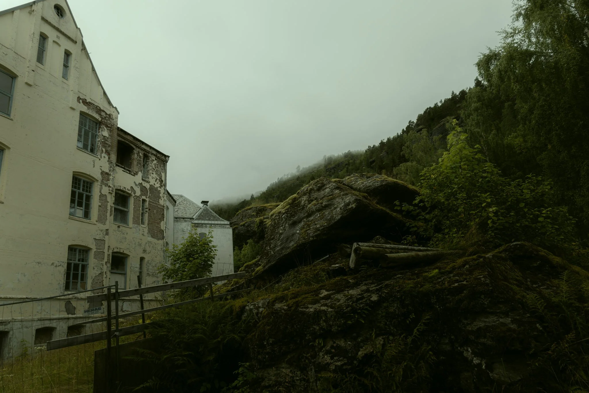 Old, weathered building on a hillside with trees and rocks, foggy mountain in the background.
