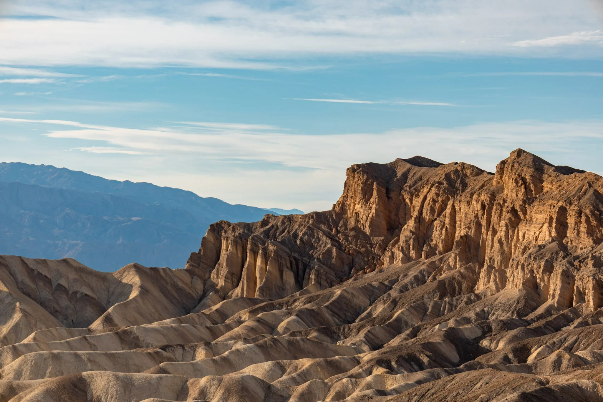 Desert landscape with layered rock formations and mountain ranges under a partly cloudy sky.