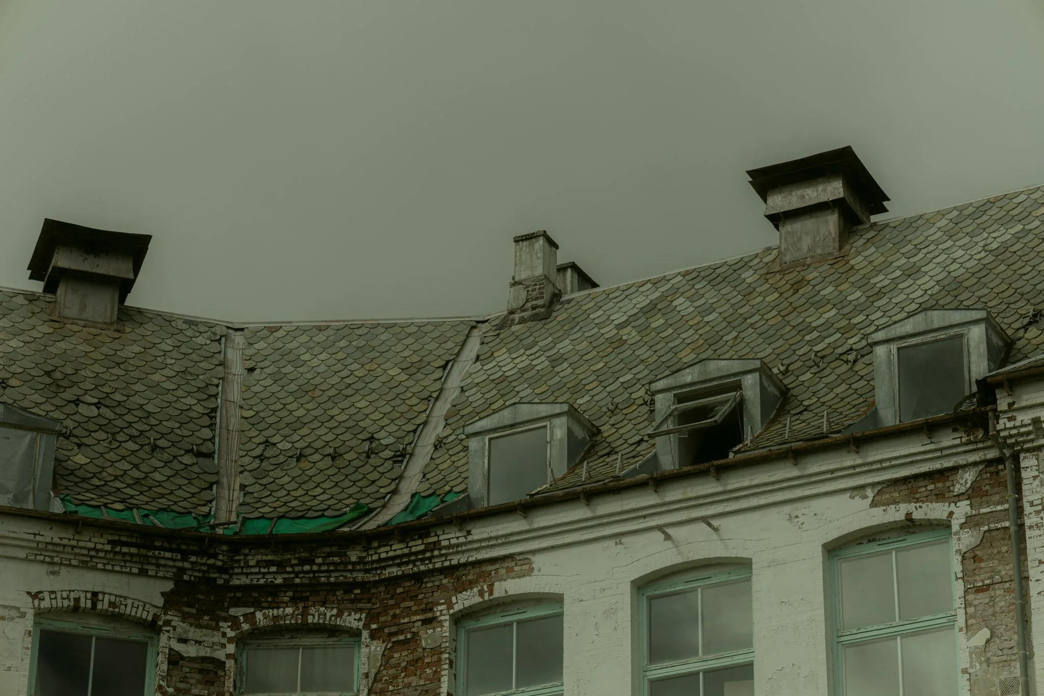 Old, deteriorated building with a sloped, shingled roof, multiple chimneys, and damaged brick and siding exterior.