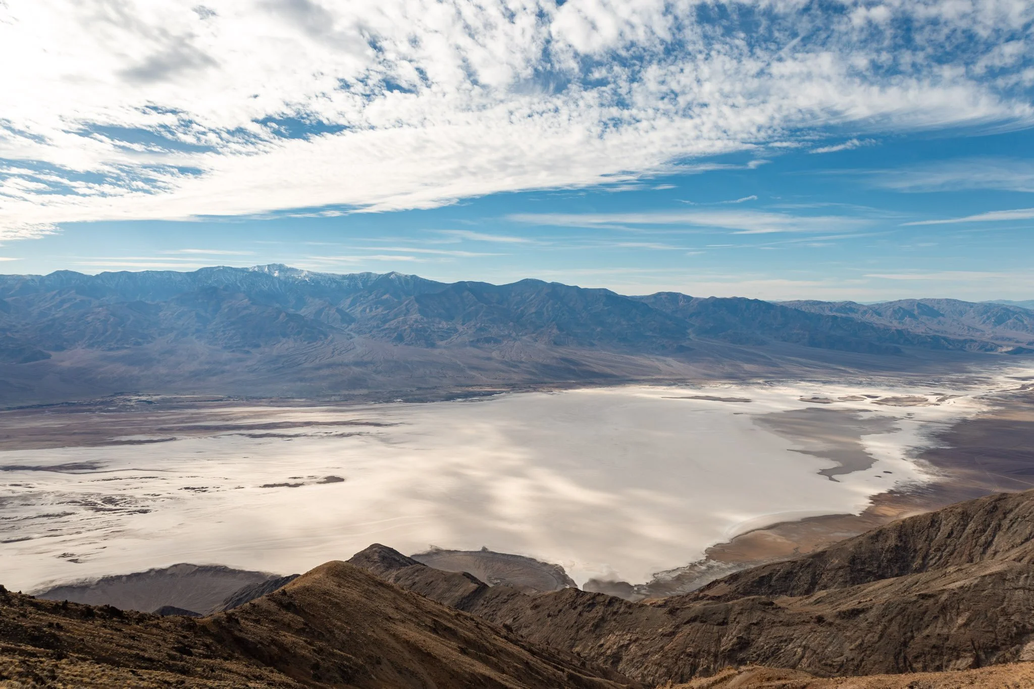 A vast landscape featuring rugged brown mountains in the foreground, a salt flat or dry lake bed in the middle, and a mountain range in the background under a partly cloudy blue sky.