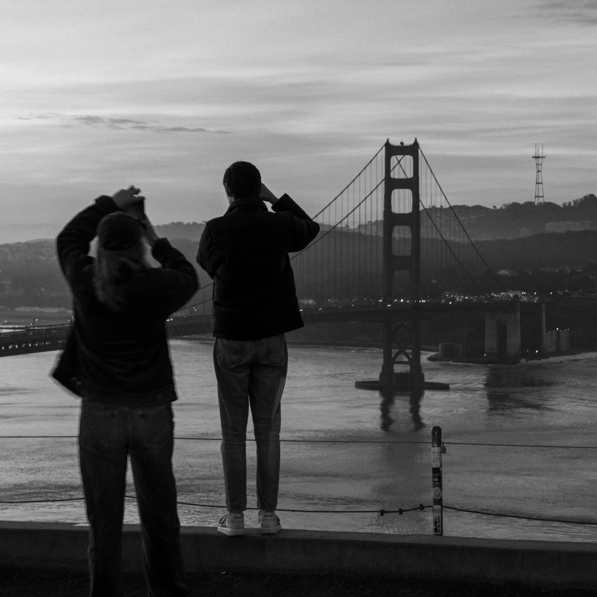 People at a waterfront with the Golden Gate Bridge in the background, some taking photos, in black and white.