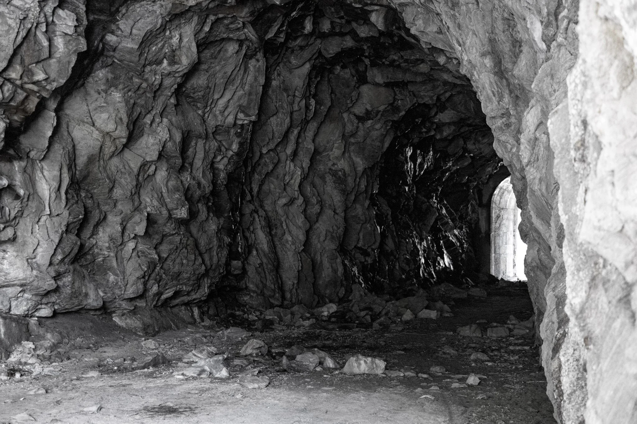 Long dark tunnel inside a rocky mountain or cave, with rough textured stone walls and ceiling, and a dirt floor.