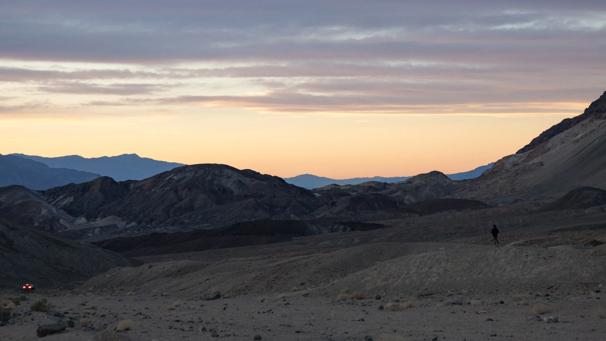 A person walking in a mountainous desert landscape at sunset or dusk with a sky filled with clouds.