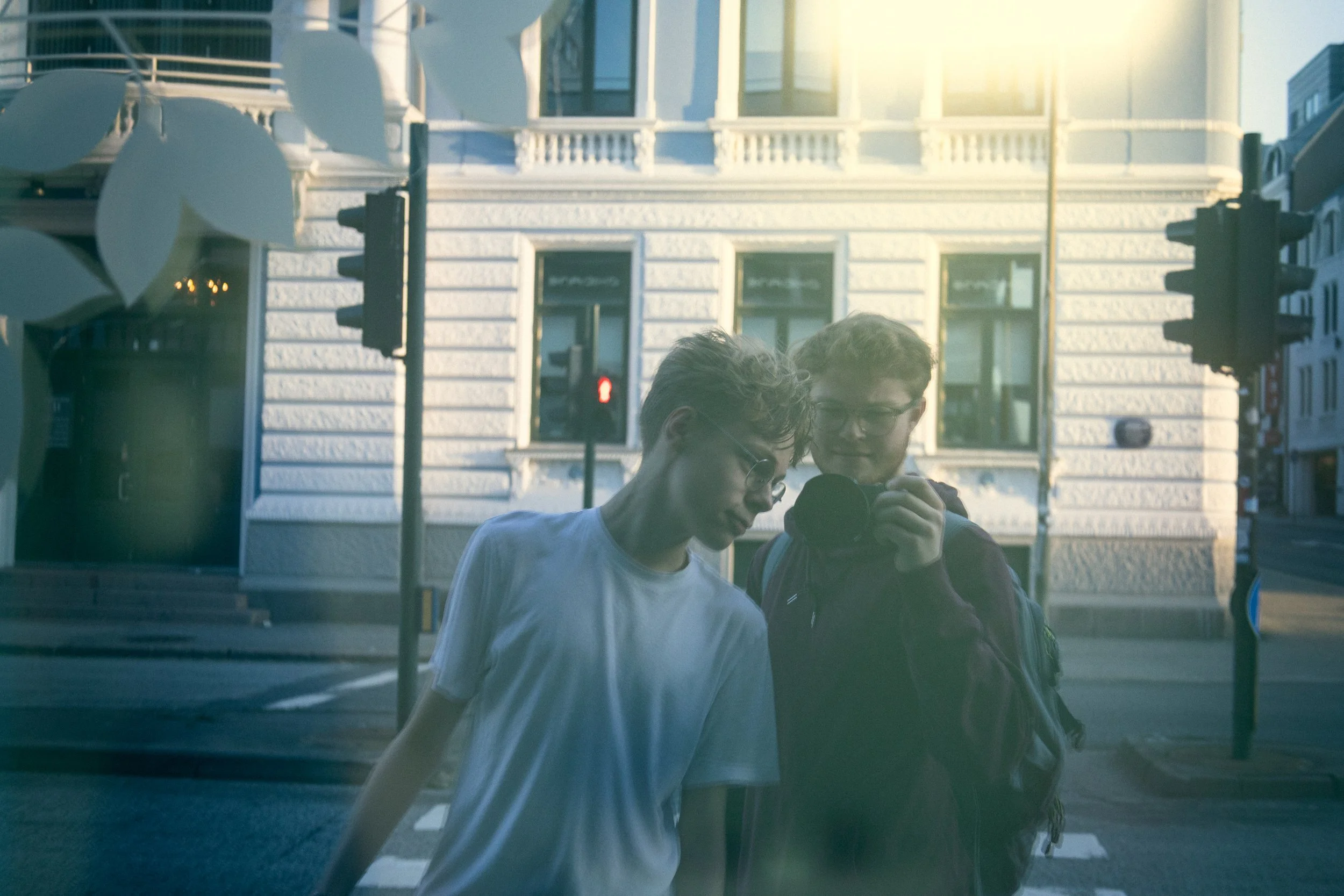 Two young men looking at a smartphone in a city street reflected in a glass surface, with urban buildings and traffic lights in the background.