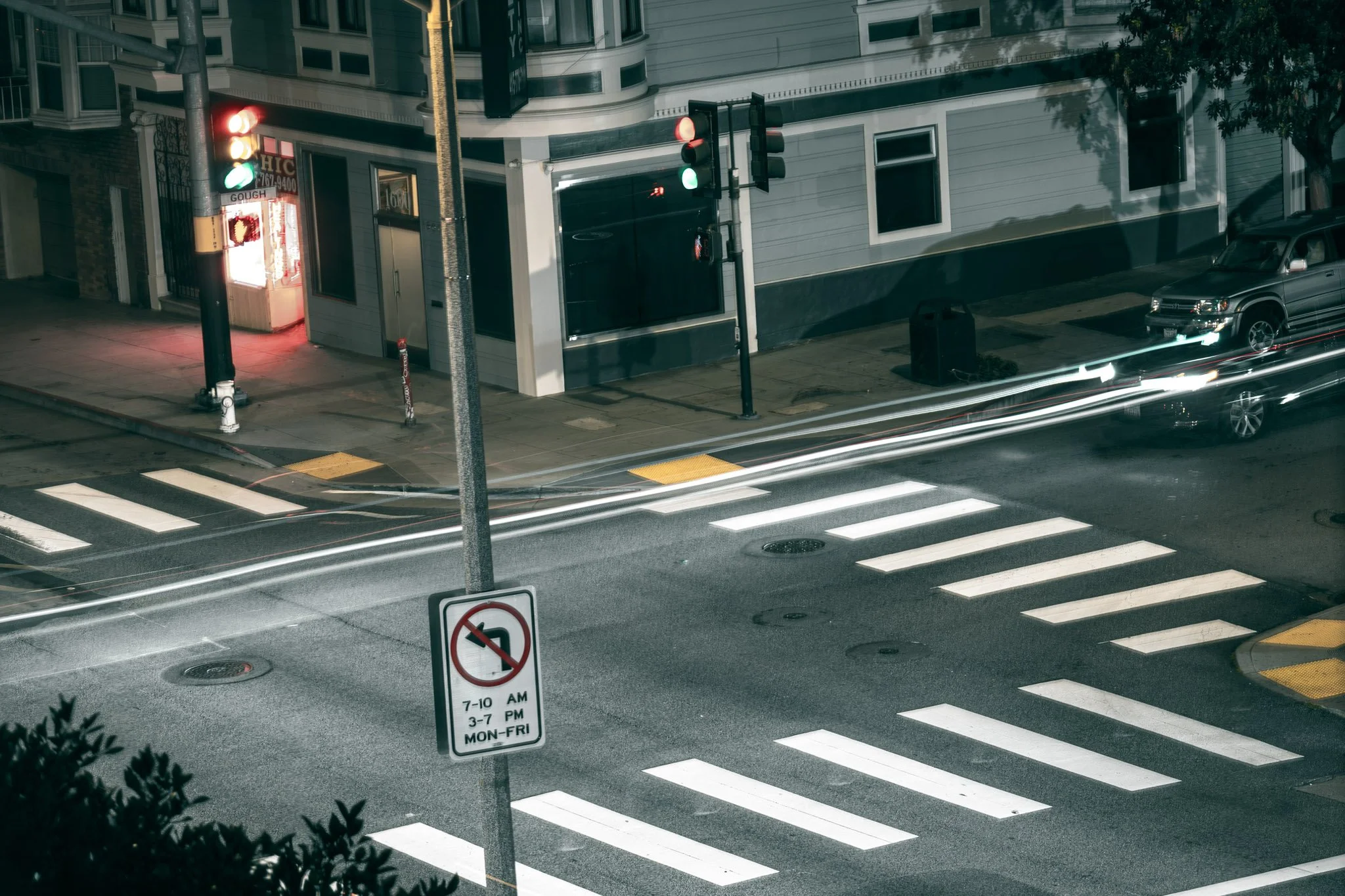 Nighttime city street scene showing a crosswalk, a traffic light, parked cars, a building with lit windows, and a street sign indicating parking restrictions.