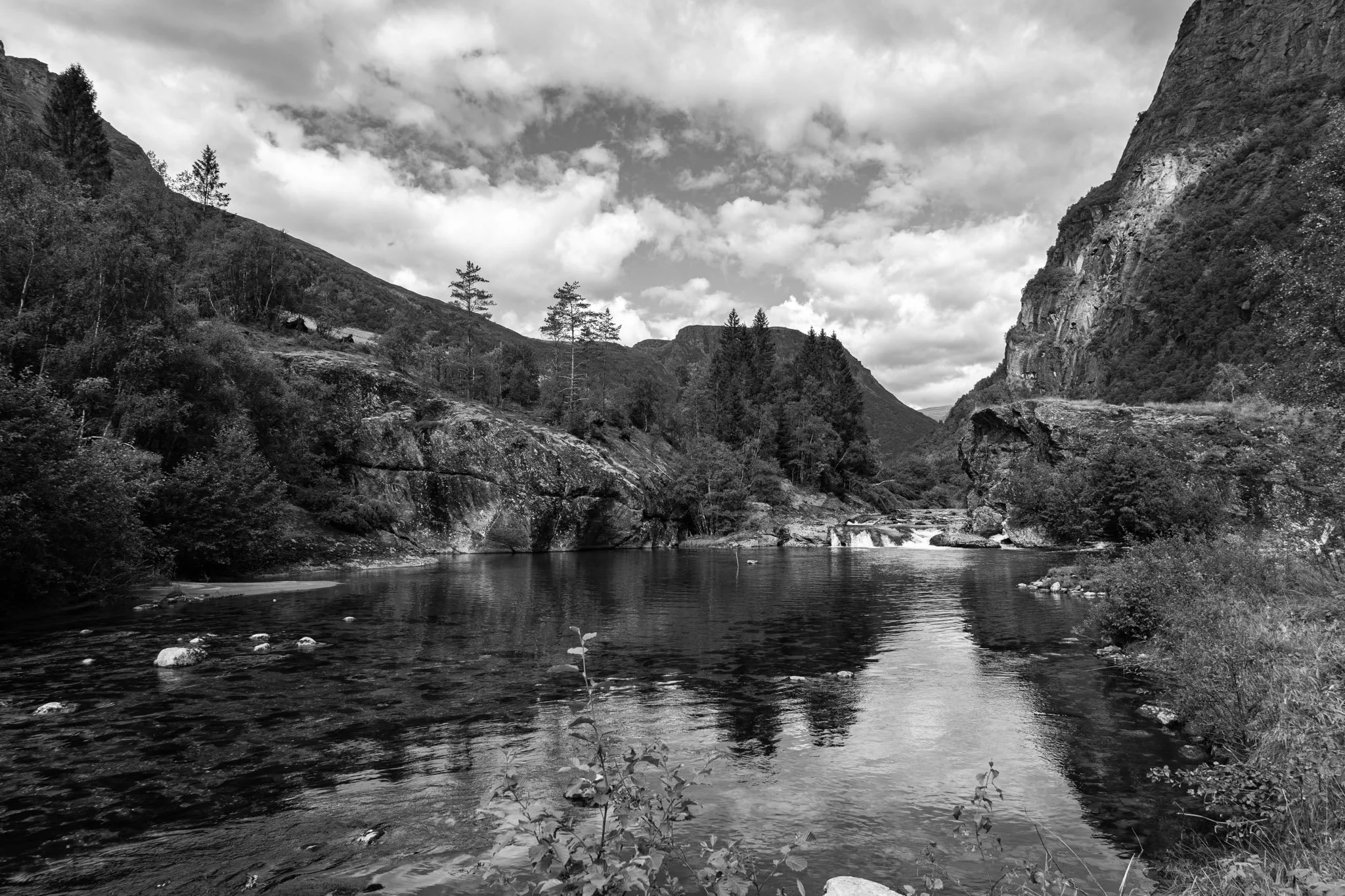 Black and white photo of a river flowing through a mountainous landscape with steep hills and a partly cloudy sky