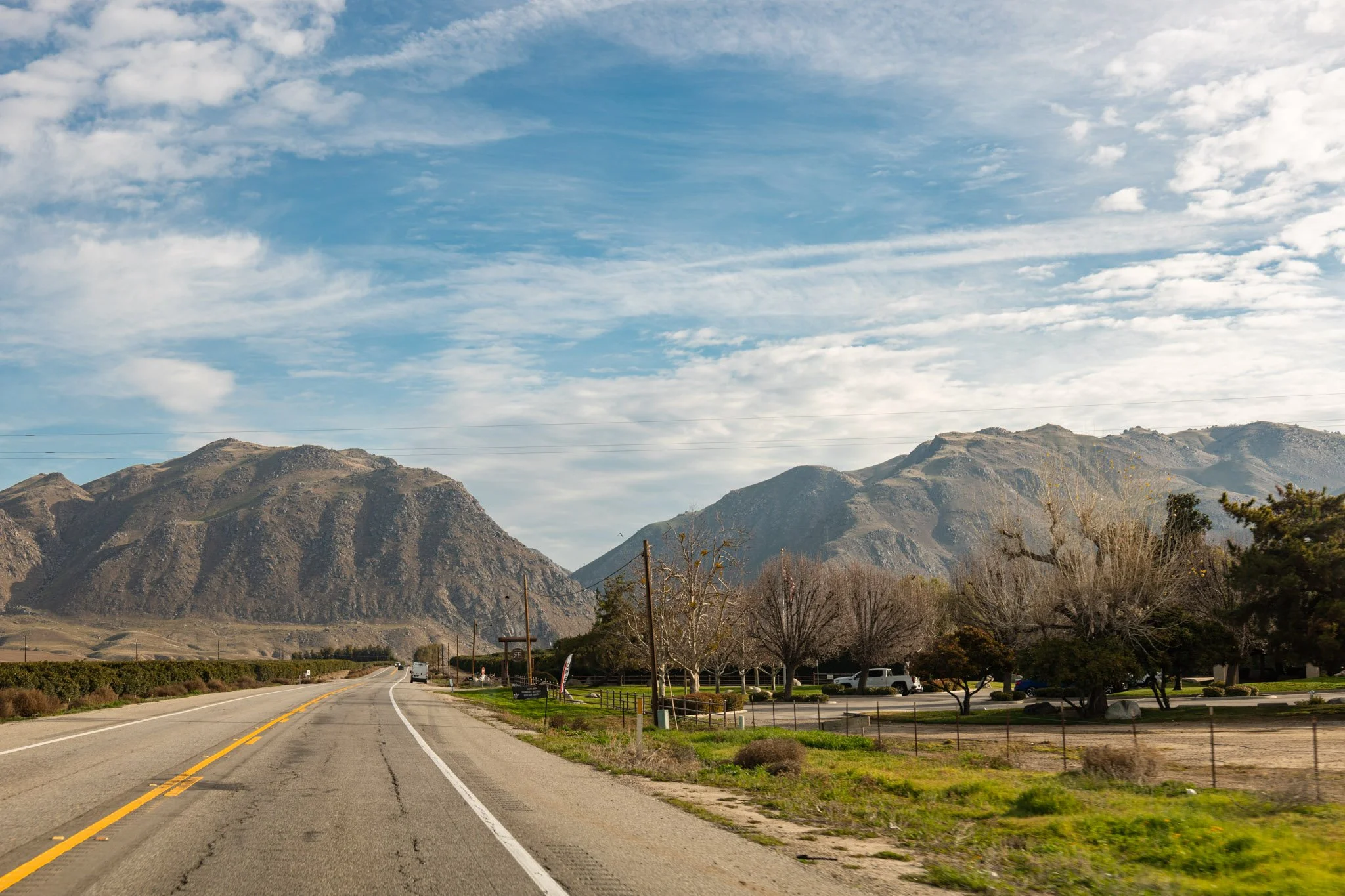 A two-lane highway leading towards mountains under a cloudy sky with patches of blue, trees, and a parking lot on the right side.