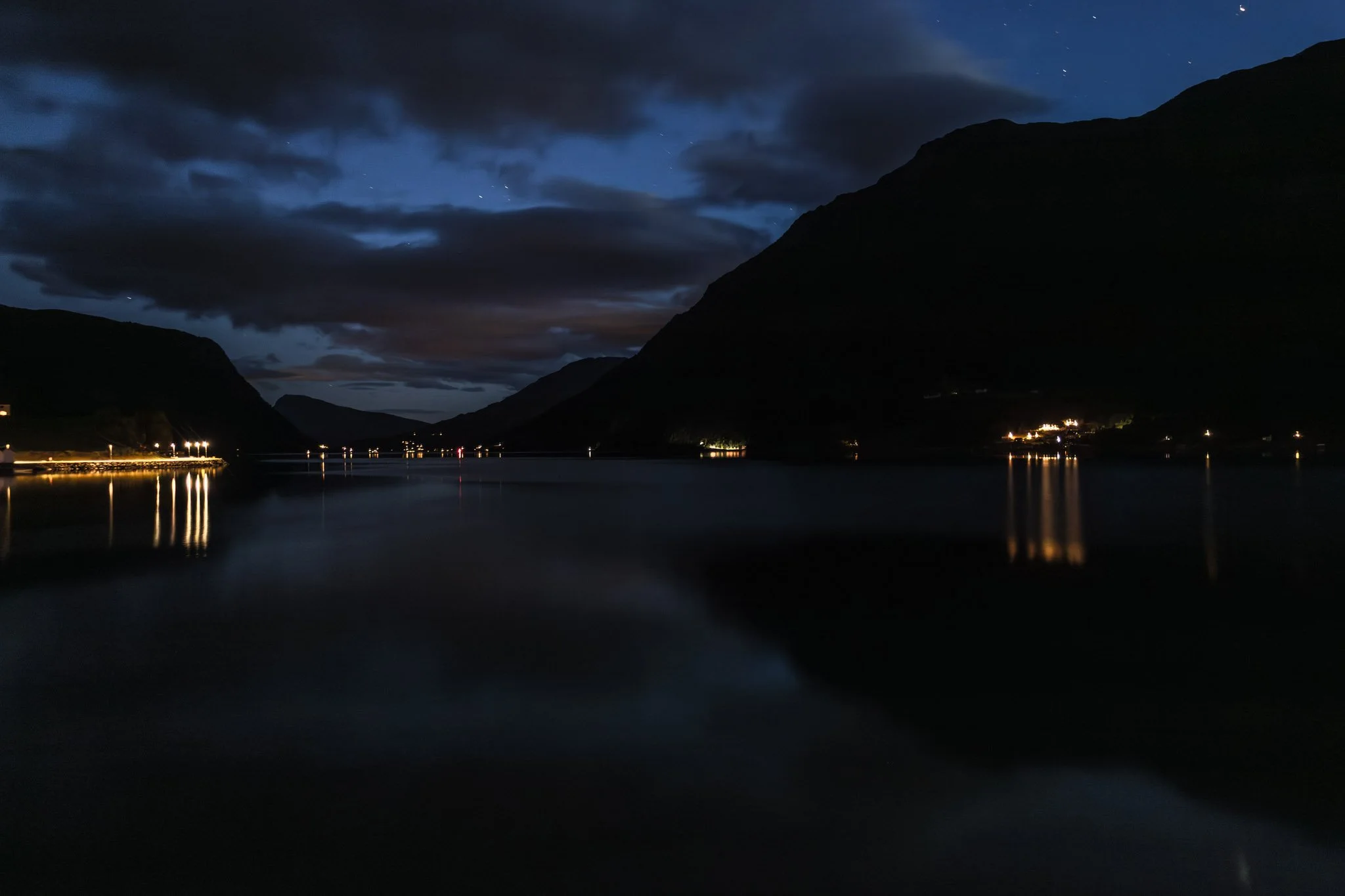 Nighttime view of a body of water surrounded by mountains with clouds overhead and small lights along the shoreline.