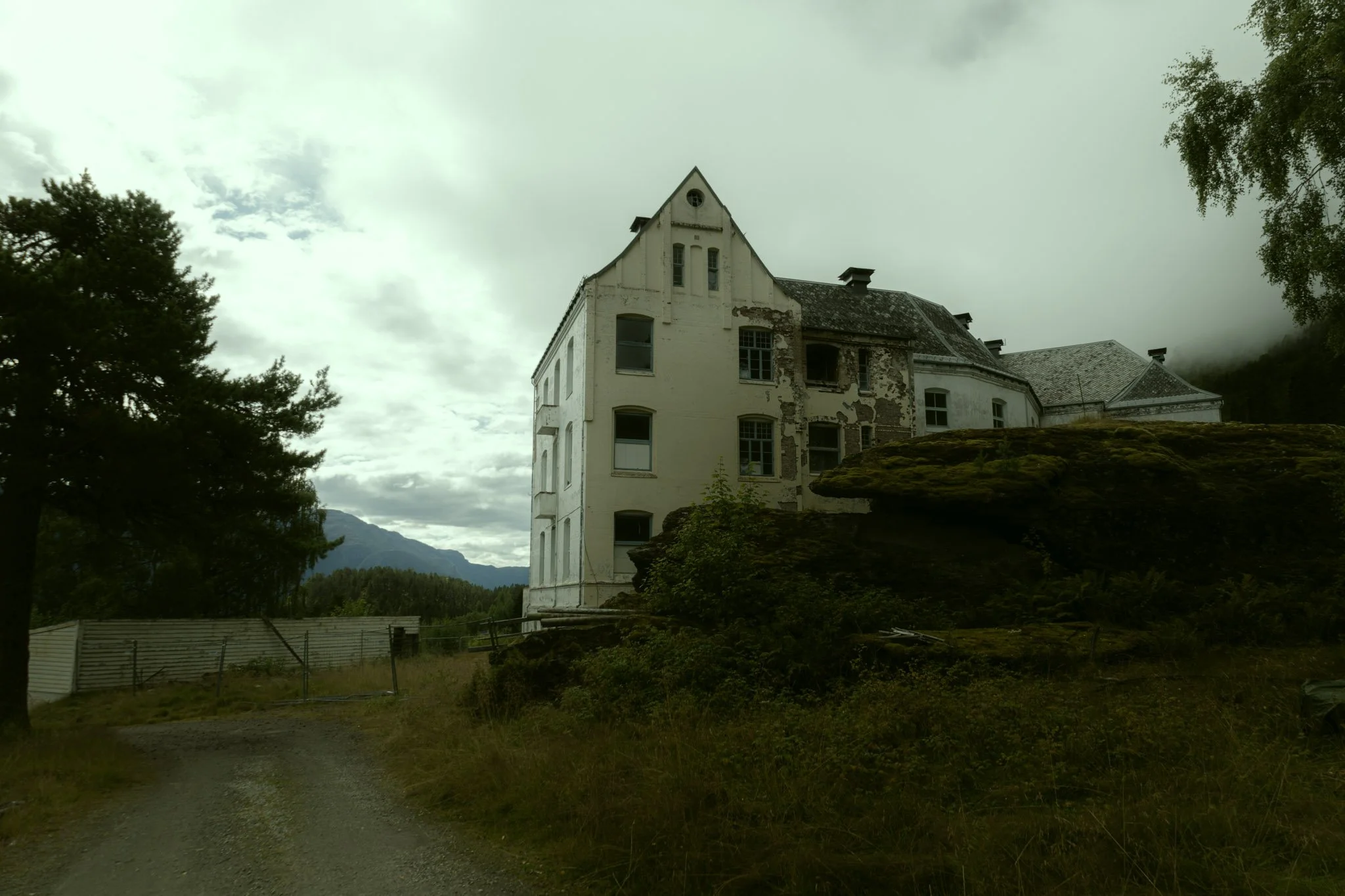 Old white house with peeling paint, surrounded by trees and mountains, under a cloudy sky.