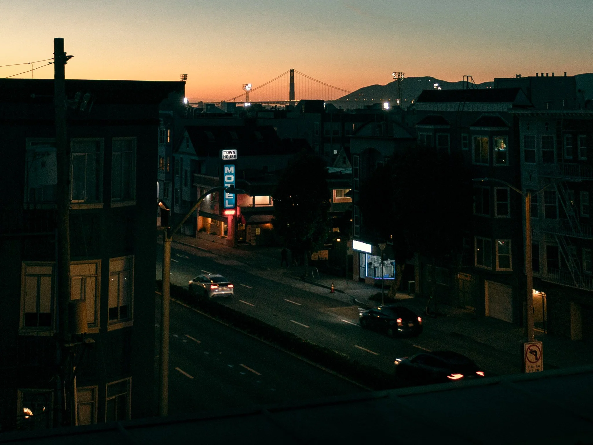 City street at dusk with buildings, cars, and traffic lights. In the background, a bridge spans a body of water with hills beyond.