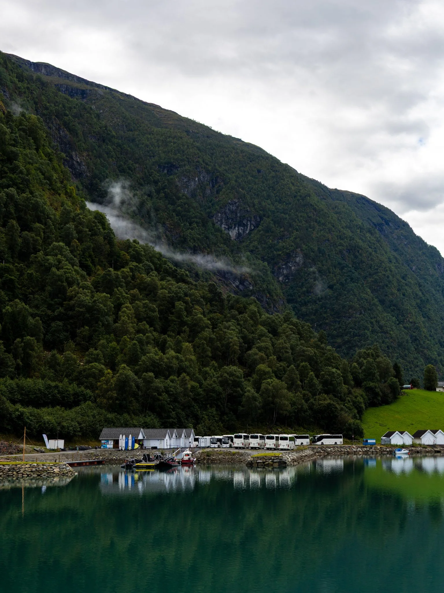 A marina with boats docked along the waterfront, surrounded by lush green mountains with a cloudy sky overhead.