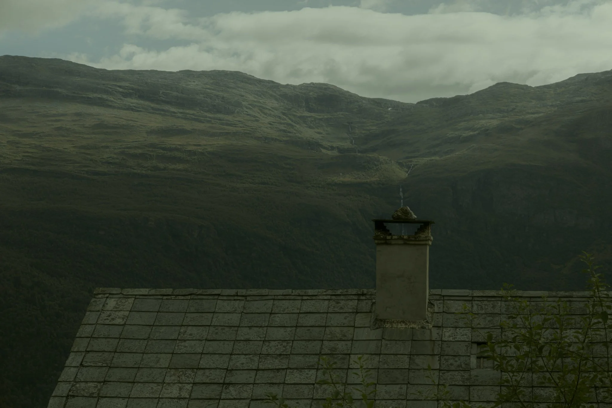 View of green, mountainous terrain with a waterfall cascading down the hillside in the distance, seen above the roof of a house with a chimney in the foreground.